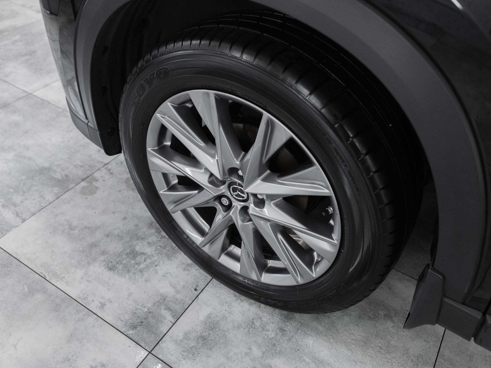 Błyszczące, szare felgi aluminiowe samochodu Mazda z oponą TOYO na tle płytek podłogowych w salonie samochodowym.