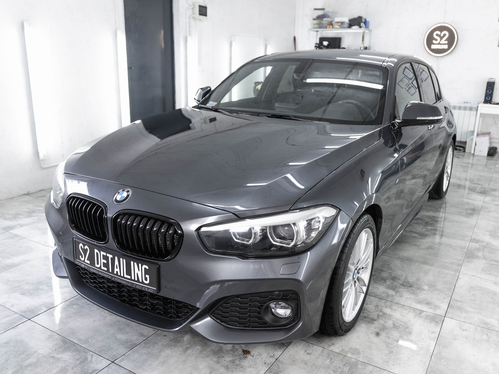 Szary samochód BMW po detailingu, z widocznym logo firmy S2 Detailing na tablicy rejestracyjnej i podświetlanym logo na ścianie warsztatu.