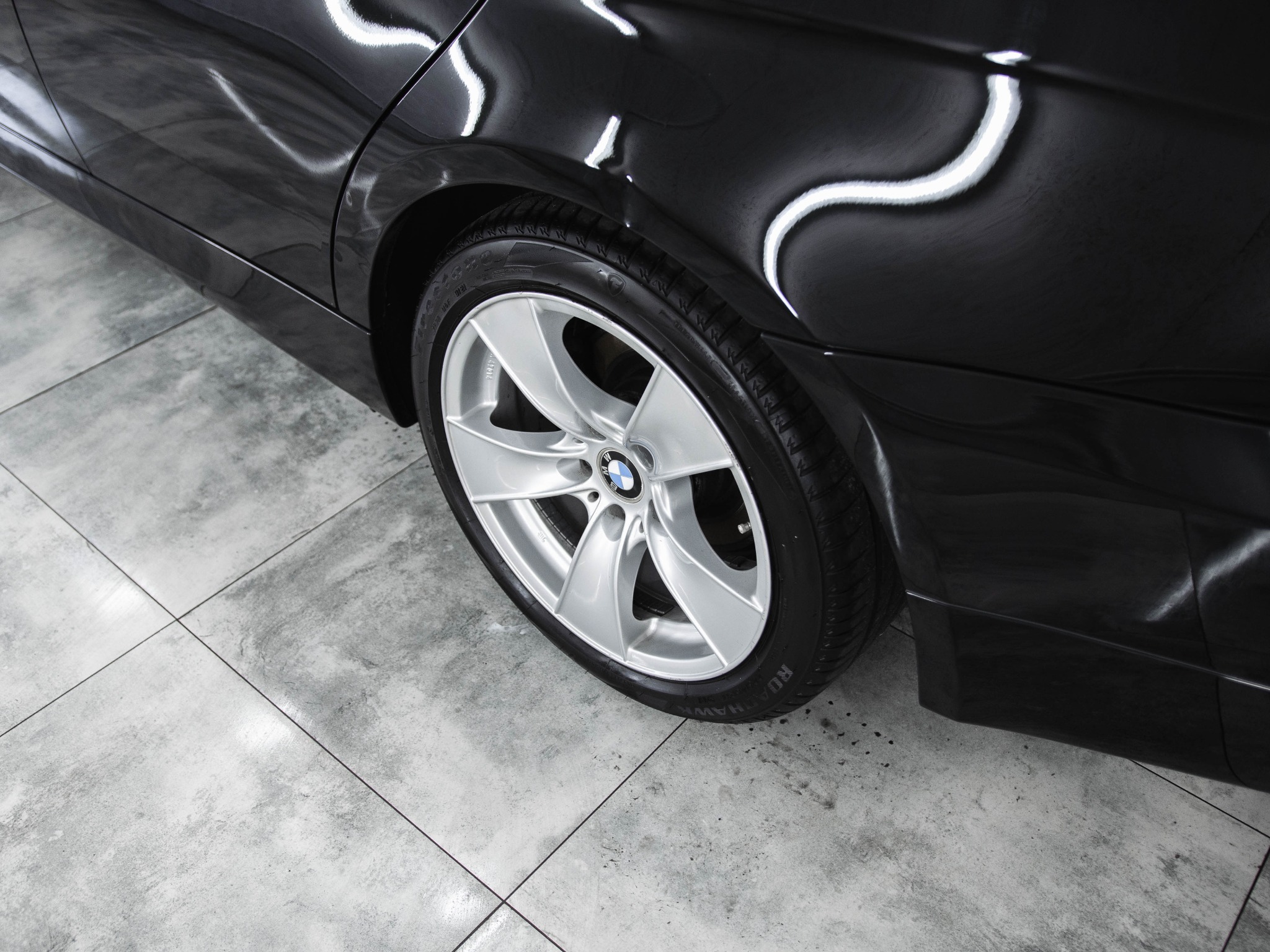 Błyszczący, czarny samochód z aluminiowymi felgami po umyciu, widoczne refleksy światła na karoserii, szare płytki podłogowe.