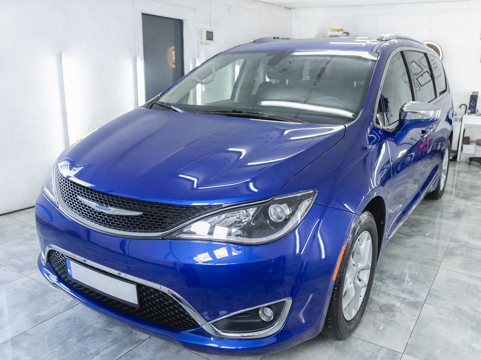 Błyszczący, granatowy Chrysler Pacifica po umyciu w myjni, widoczny z przodu, z refleksami światła na karoserii i czystymi felgami, w jasnym pomieszczeniu z białymi ścianami i szarą podłogą.