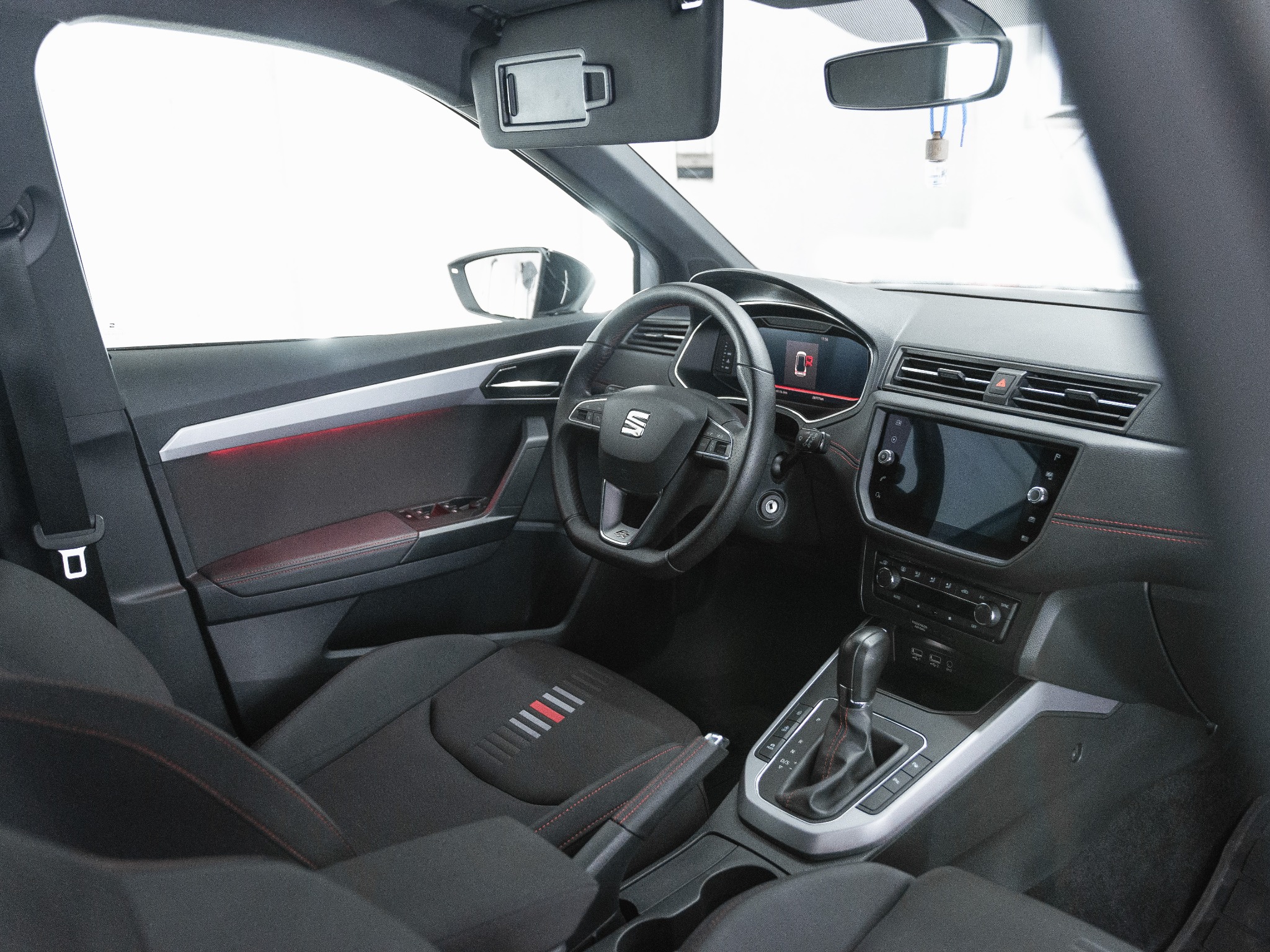 Wnętrze samochodu marki Seat z czarną tapicerką i czerwonymi akcentami, widoczna deska rozdzielcza, kierownica z logo, lewarek zmiany biegów i fragment fotela kierowcy z pasami w kolorach...