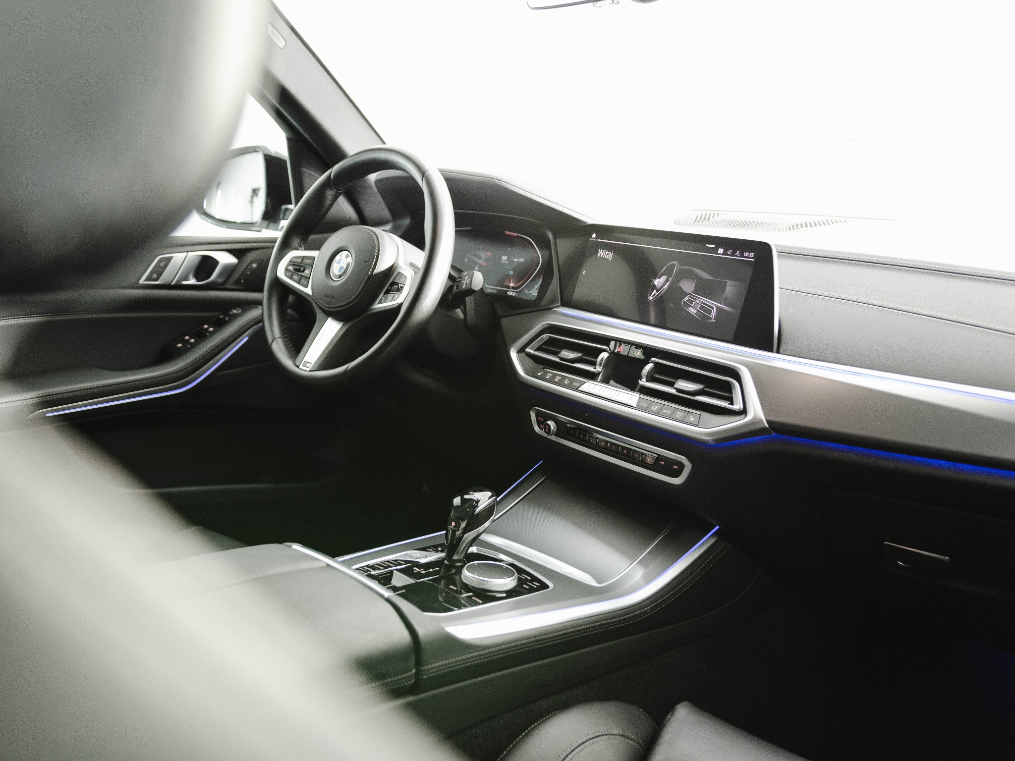 Wnętrze samochodu BMW X5 z widocznym kokpitem, kierownicą z logo, ekranem dotykowym i podświetleniem ambientowym w kolorze niebieskim.
