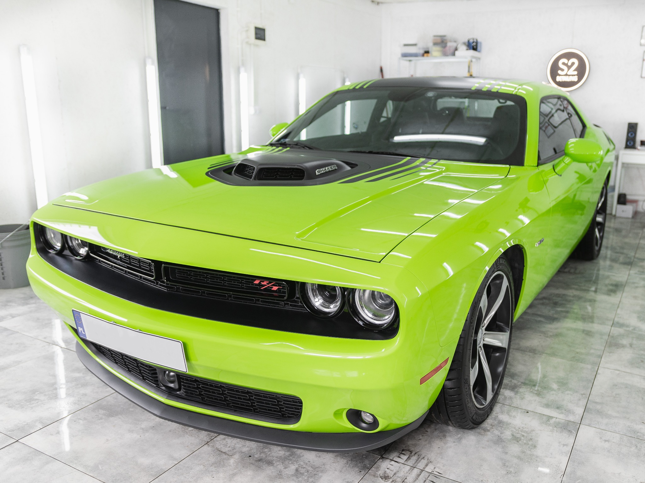 Jaskrawozielony Dodge Challenger z czarnymi pasami na masce i dachu, zaparkowany w jasnym, sterylnym pomieszczeniu z błyszczącą podłogą z płytek. Widoczny logotyp 'S2 Detailing' w tle.