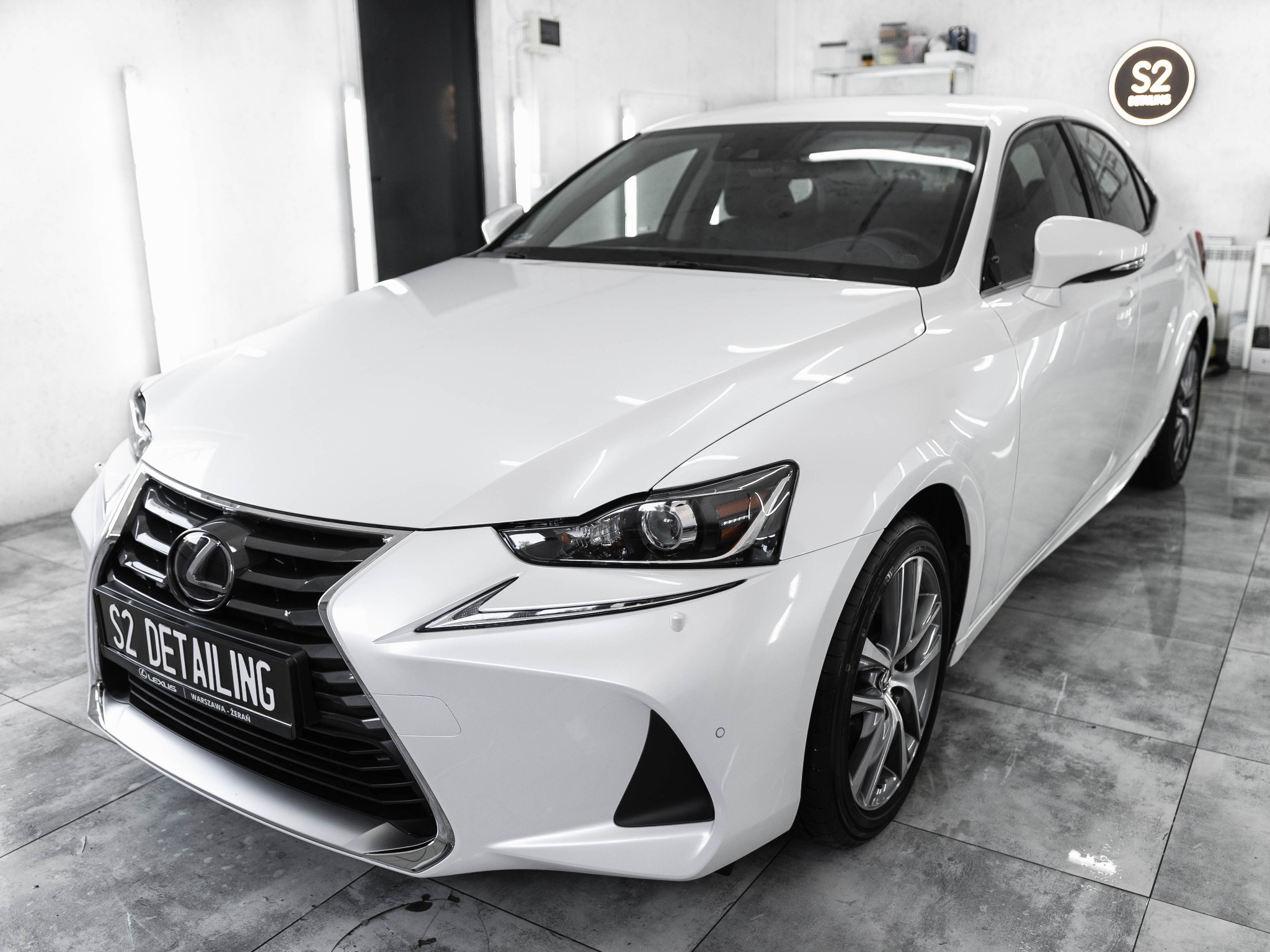 Biały Lexus IS po detailingu, widoczny z przodu w jasnym pomieszczeniu z logo S2 Detailing w tle.