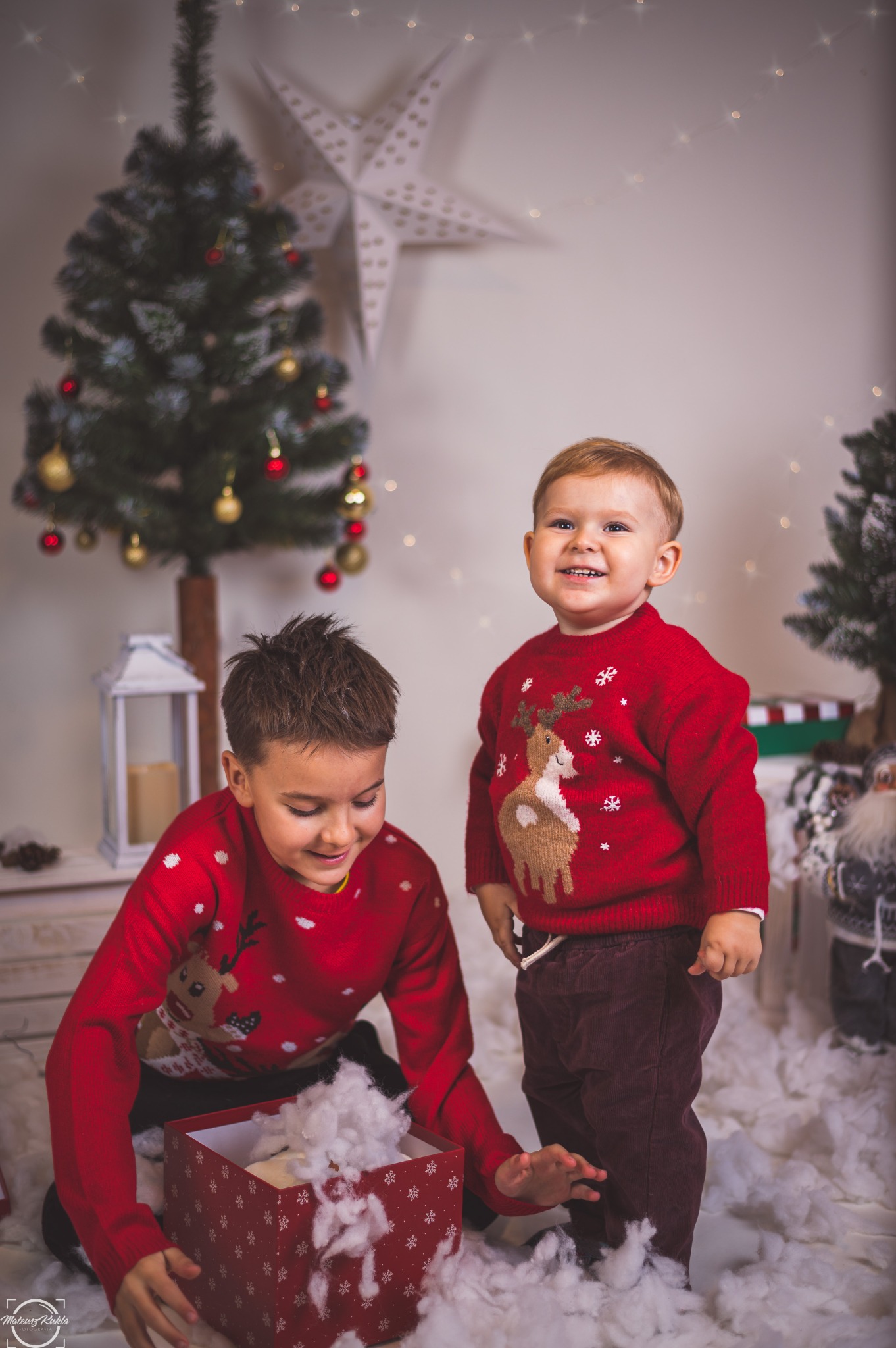Dwóch chłopców w czerwonych świątecznych swetrach bawi się w zimowej scenerii z choinką i dekoracjami, jeden wyciąga watę z pudełka.