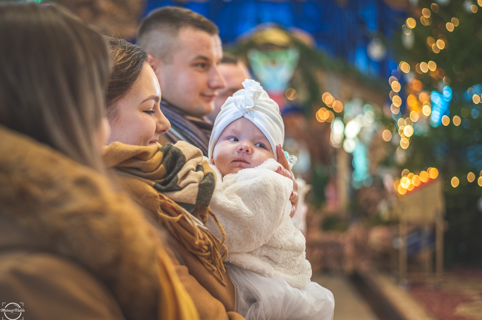 Uśmiechnięta matka trzymająca niemowlę w białym ubranku i turbanie na tle kościoła z choinką