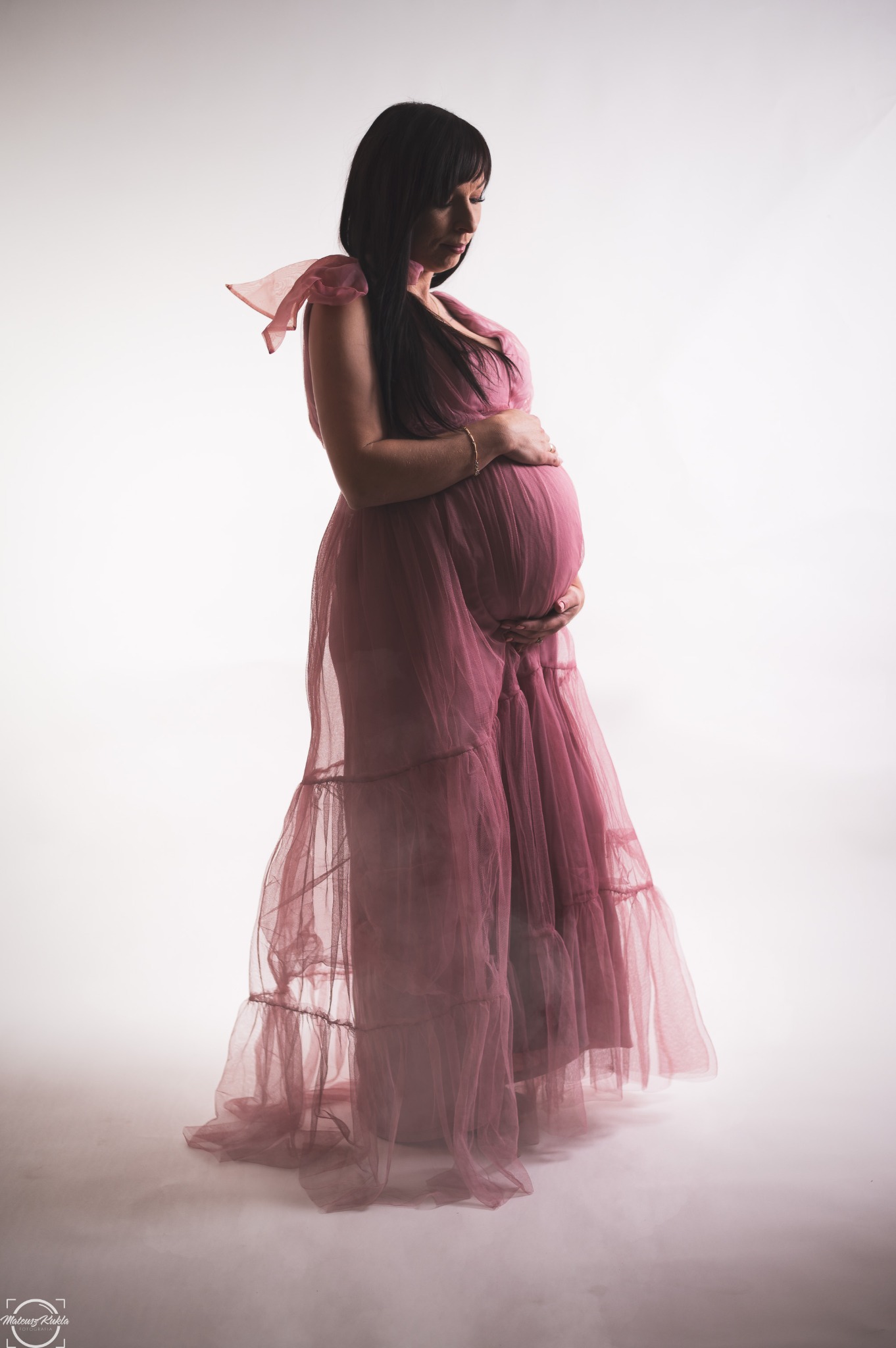 Kobieta w zwiewnej, różowej sukni ciążowej, delikatnie obejmująca brzuch na jasnym tle, z widocznym złotym pierścionkiem i bransoletką.