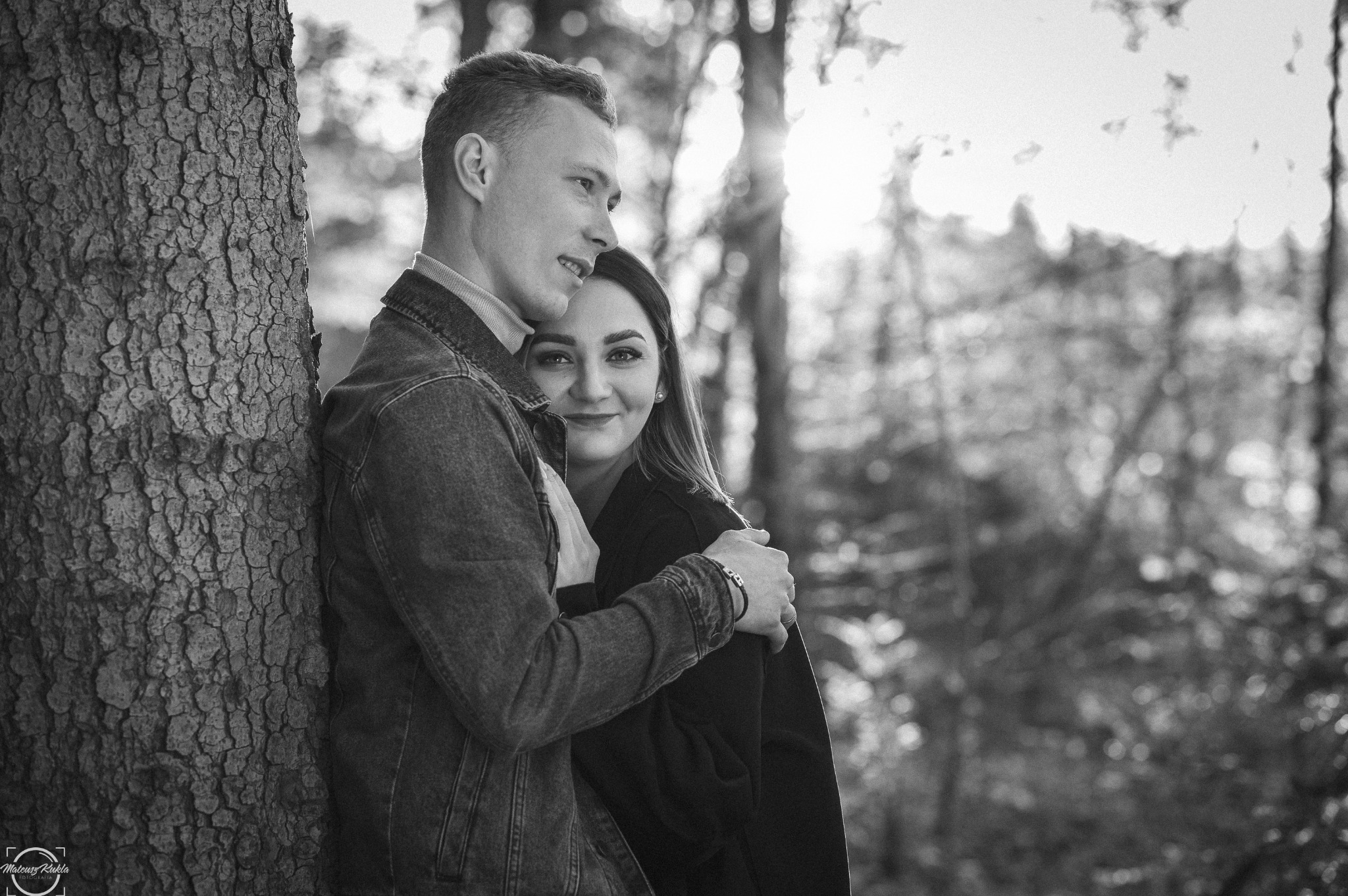 Czarno-białe zdjęcie pary zakochanych obejmujących się przy drzewie w lesie, z delikatnym uśmiechem kobiety skierowanym w stronę obiektywu, mężczyzna oparty o korę drzewa w denimowej kurtce.