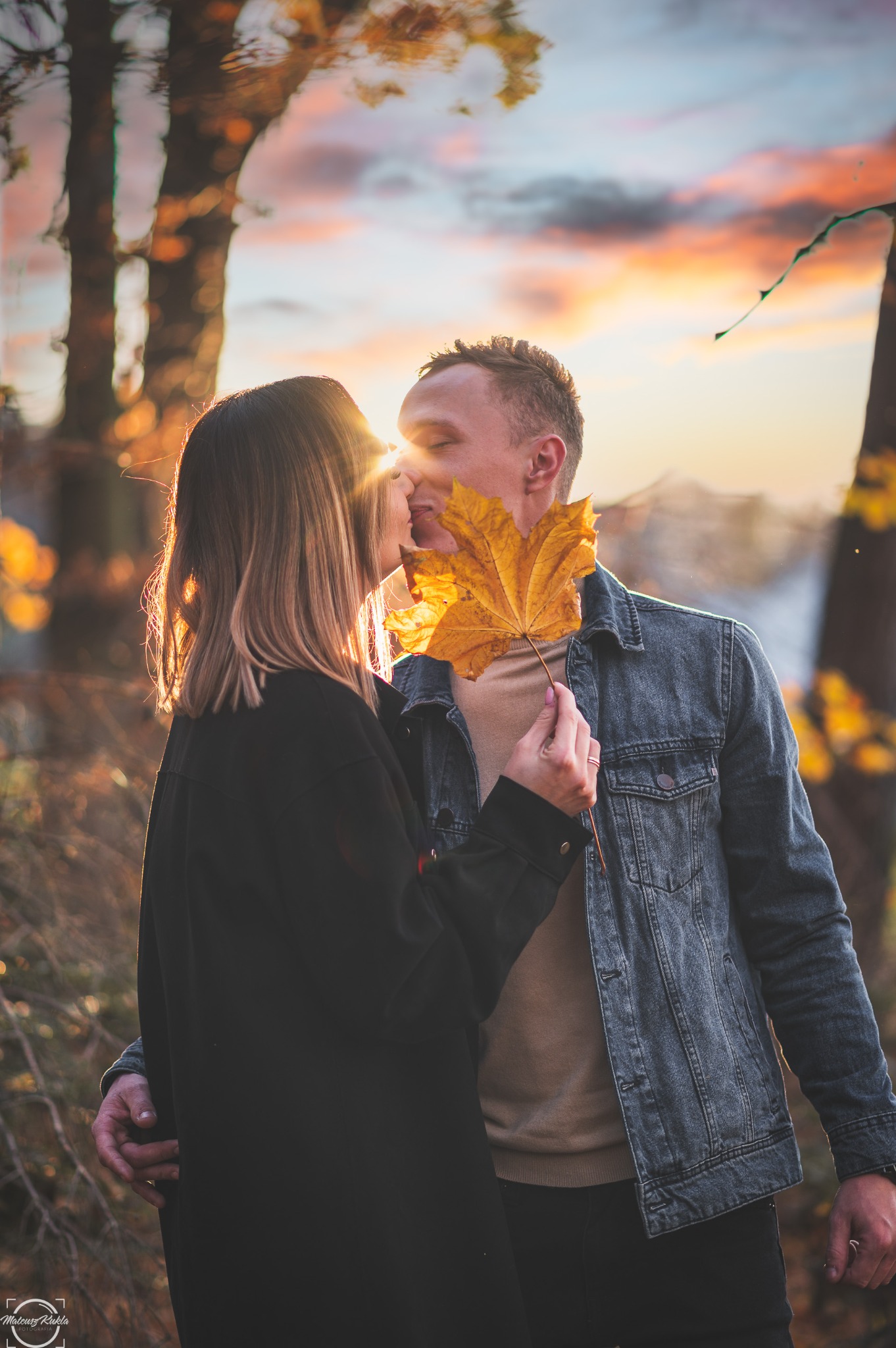 Zakochana para całuje się na tle jesiennego zachodu słońca, trzymając przed sobą duży, żółty liść klonu, który częściowo zasłania ich twarze.