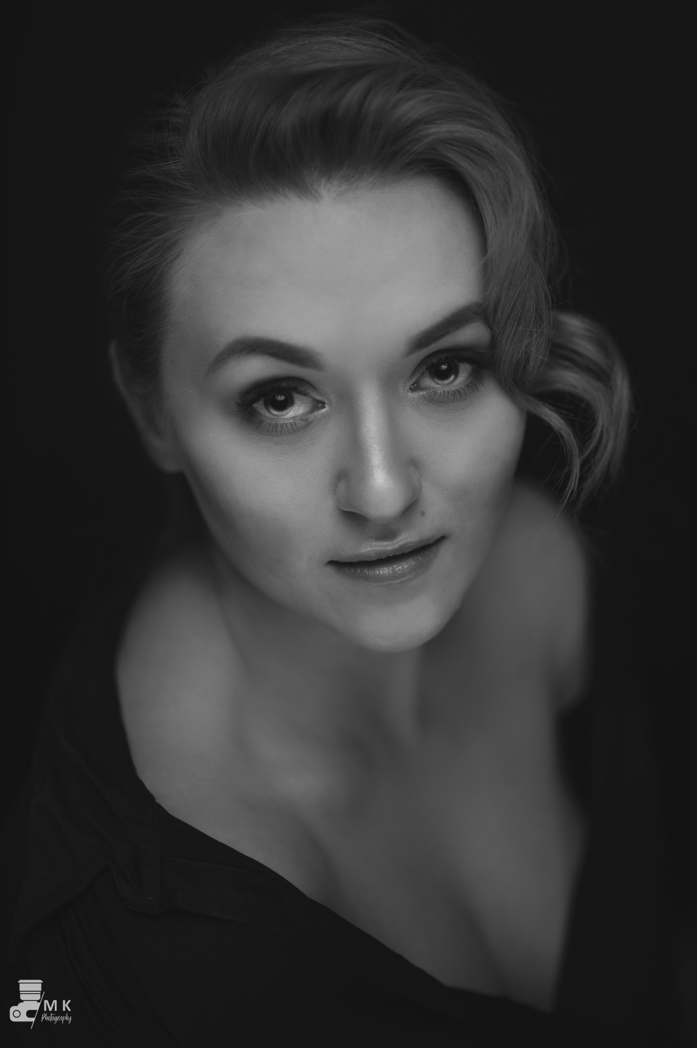 Portret kobiety w czerni i bieli, z delikatnym makijażem i upiętymi włosami, spoglądającej w górę na obiektyw.
