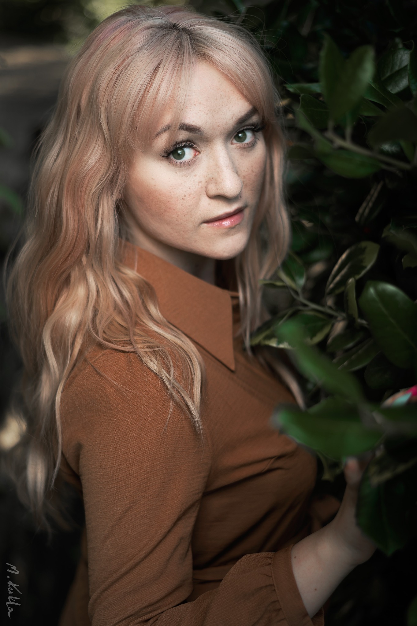 Portret kobiety z piegami i blond włosami w brązowej koszuli na tle zielonych liści.