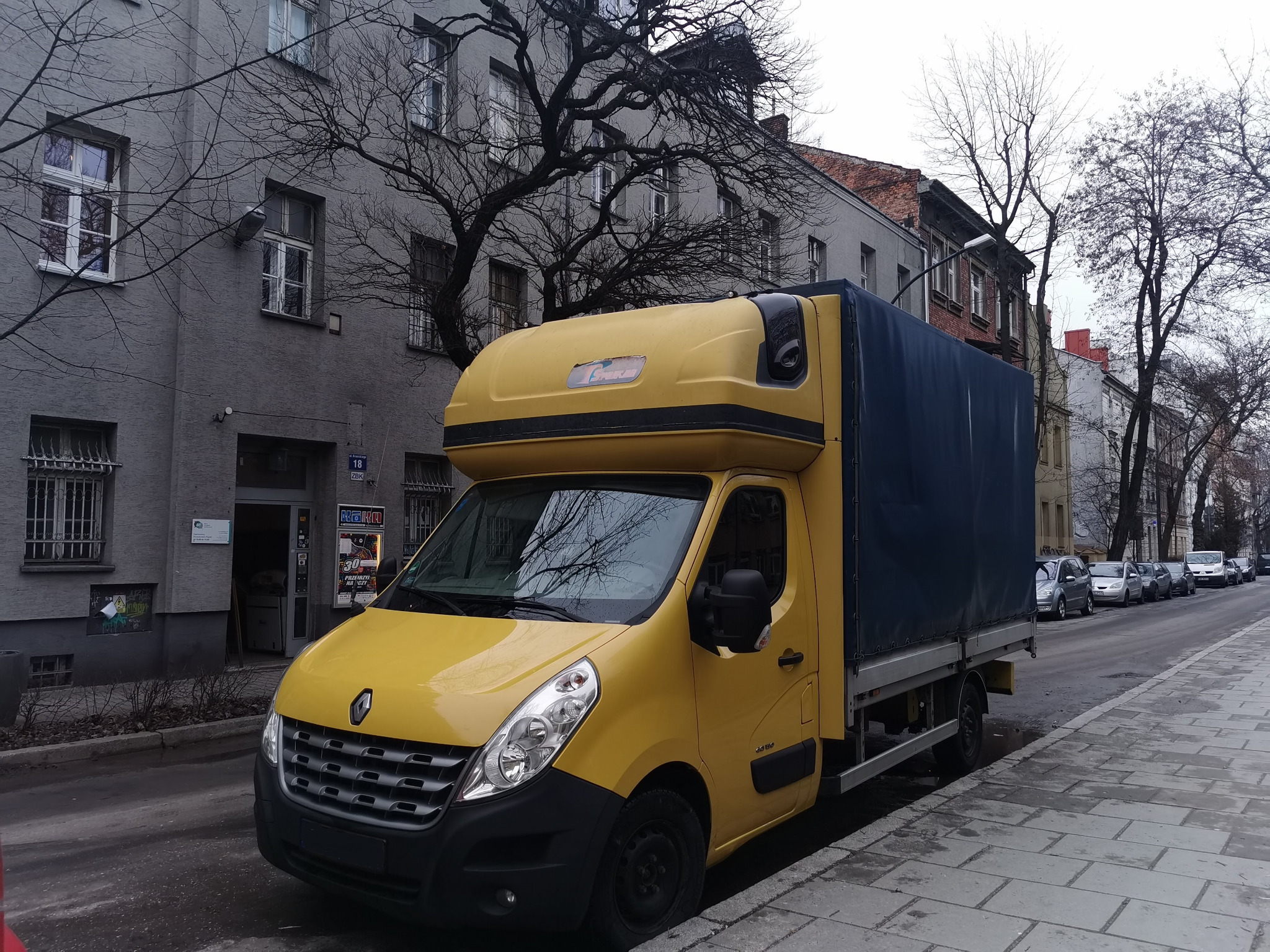 Żółty samochód dostawczy Renault Master z plandeką na parkingu miejskim, widoczne budynki w tle i drzewa bez liści.