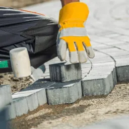 Pracownik w żółto-szarych rękawicach układa szarą kostkę brukową o nieregularnym kształcie, tworząc łukowaty wzór; obok leży gumowy młotek.