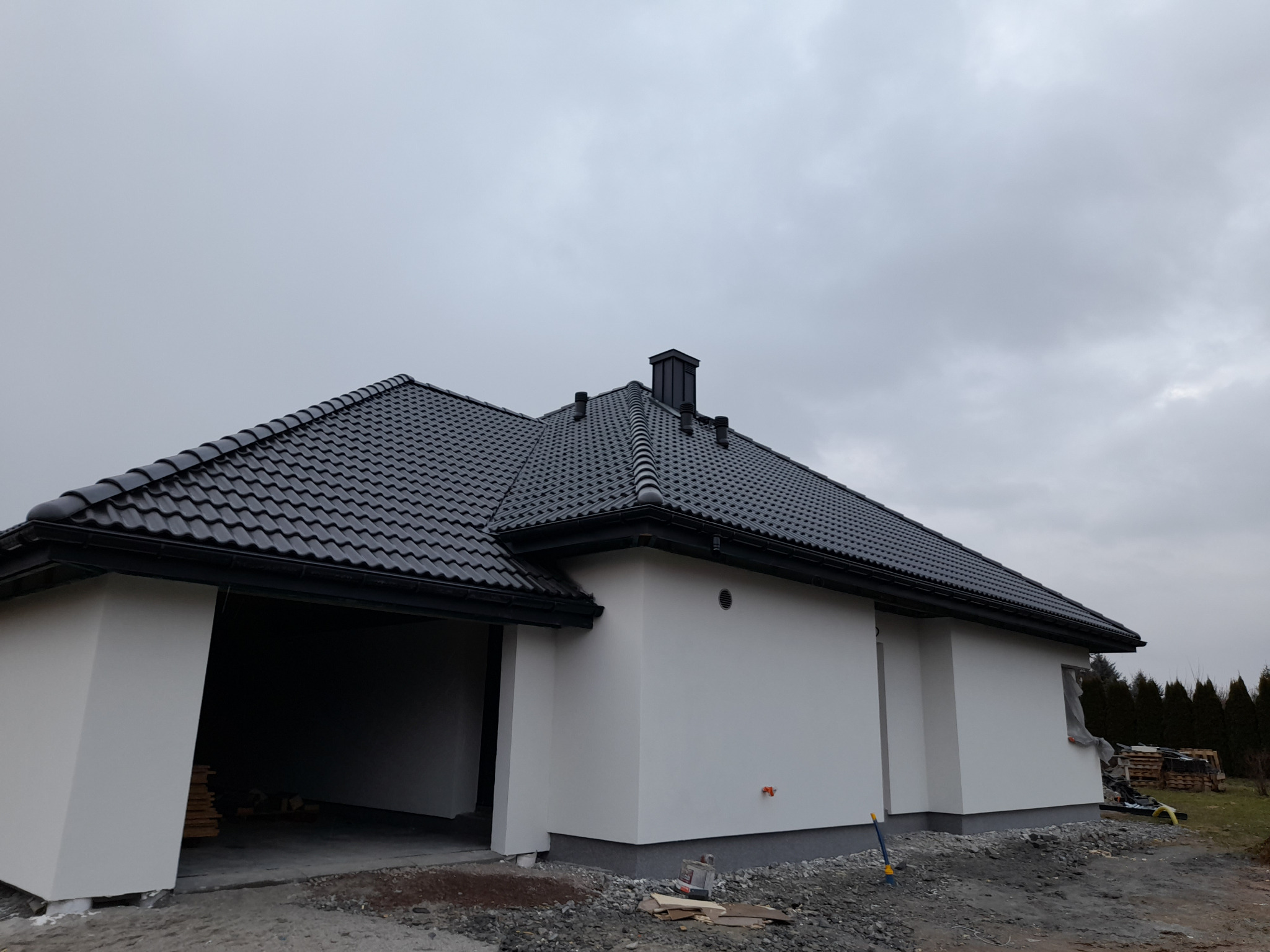 Nowoczesny dom jednorodzinny z ciemną dachówką ceramiczną, widoczne rynny i komin, biała elewacja, teren wokół domu w trakcie prac wykończeniowych, pochmurne niebo.