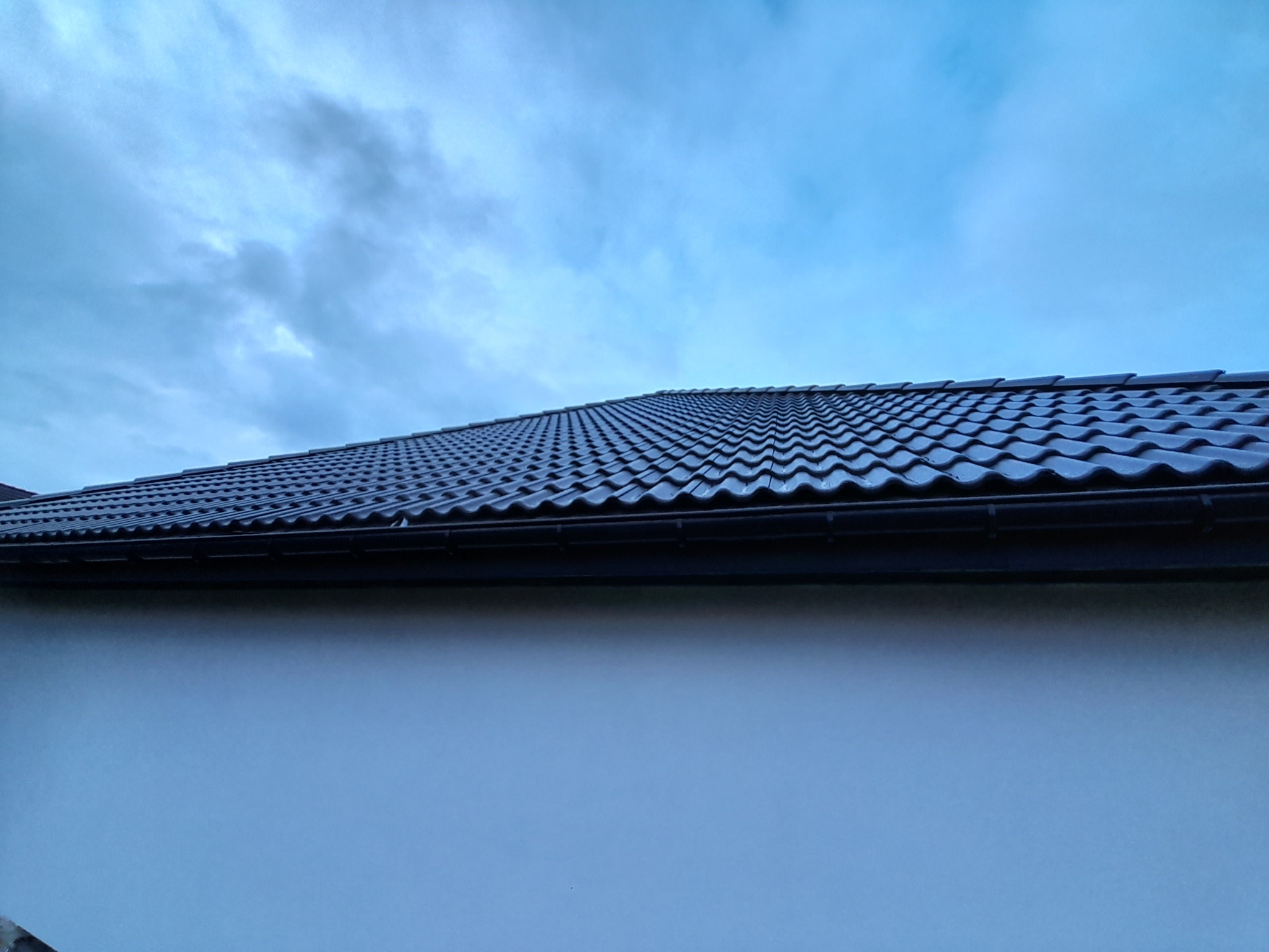 Nowy, ciemny dach z dachówki ceramicznej, widok z dołu na tle pochmurnego nieba, z fragmentem ściany budynku.