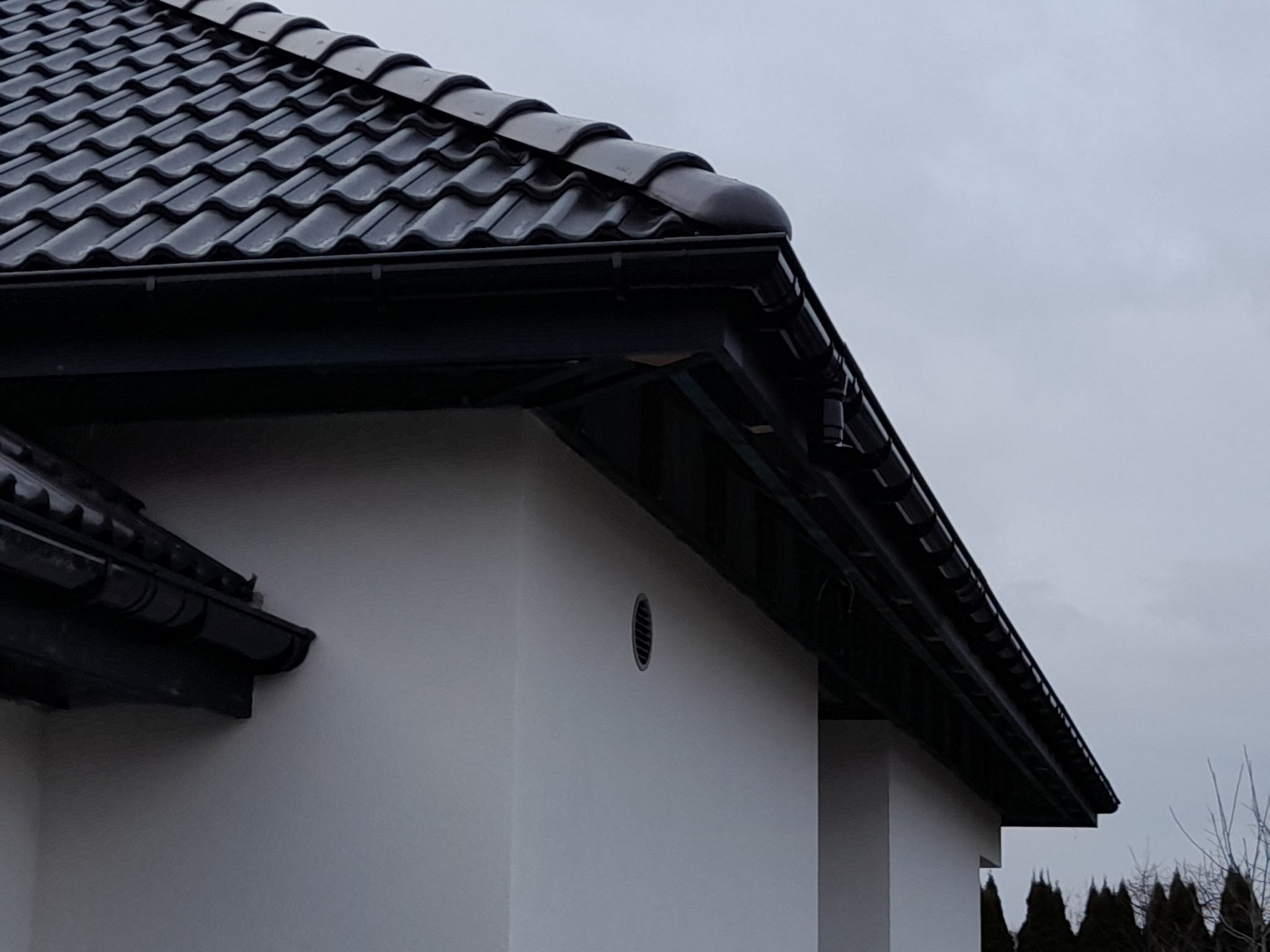 Fragment dachu z czarną dachówką ceramiczną i systemem rynnowym w kolorze antracytu, widok z dołu na tle pochmurnego nieba.