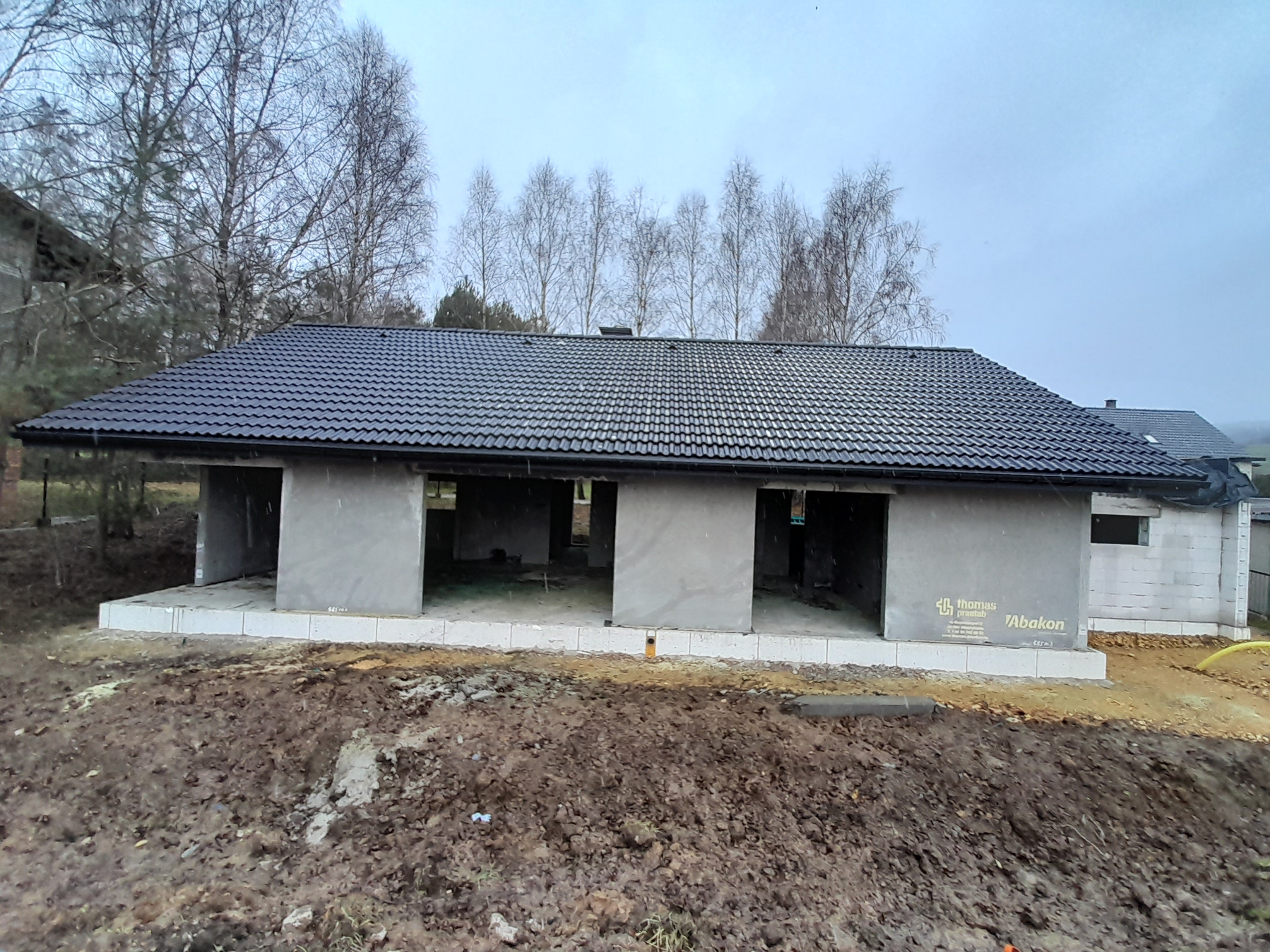 Surowy budynek w trakcie budowy z nowym, ciemnym pokryciem na dachu, widoczne drzewa w tle, pochmurne niebo.