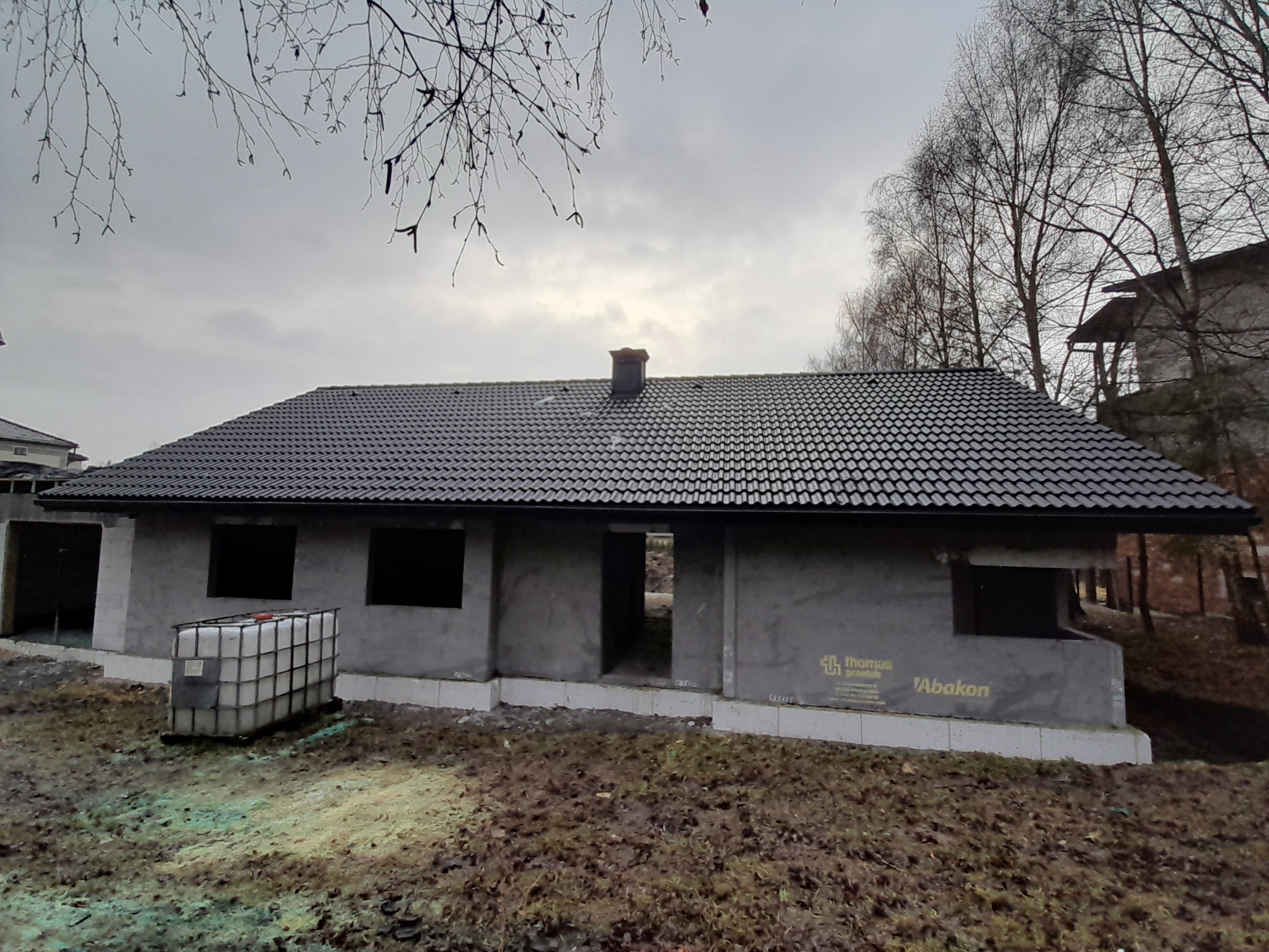 Nowo położony, ciemny dach z dachówki ceramicznej na surowym budynku w stanie deweloperskim, widoczny komin i zbiornik na wodę, jesienne drzewa w tle pod pochmurnym niebem.