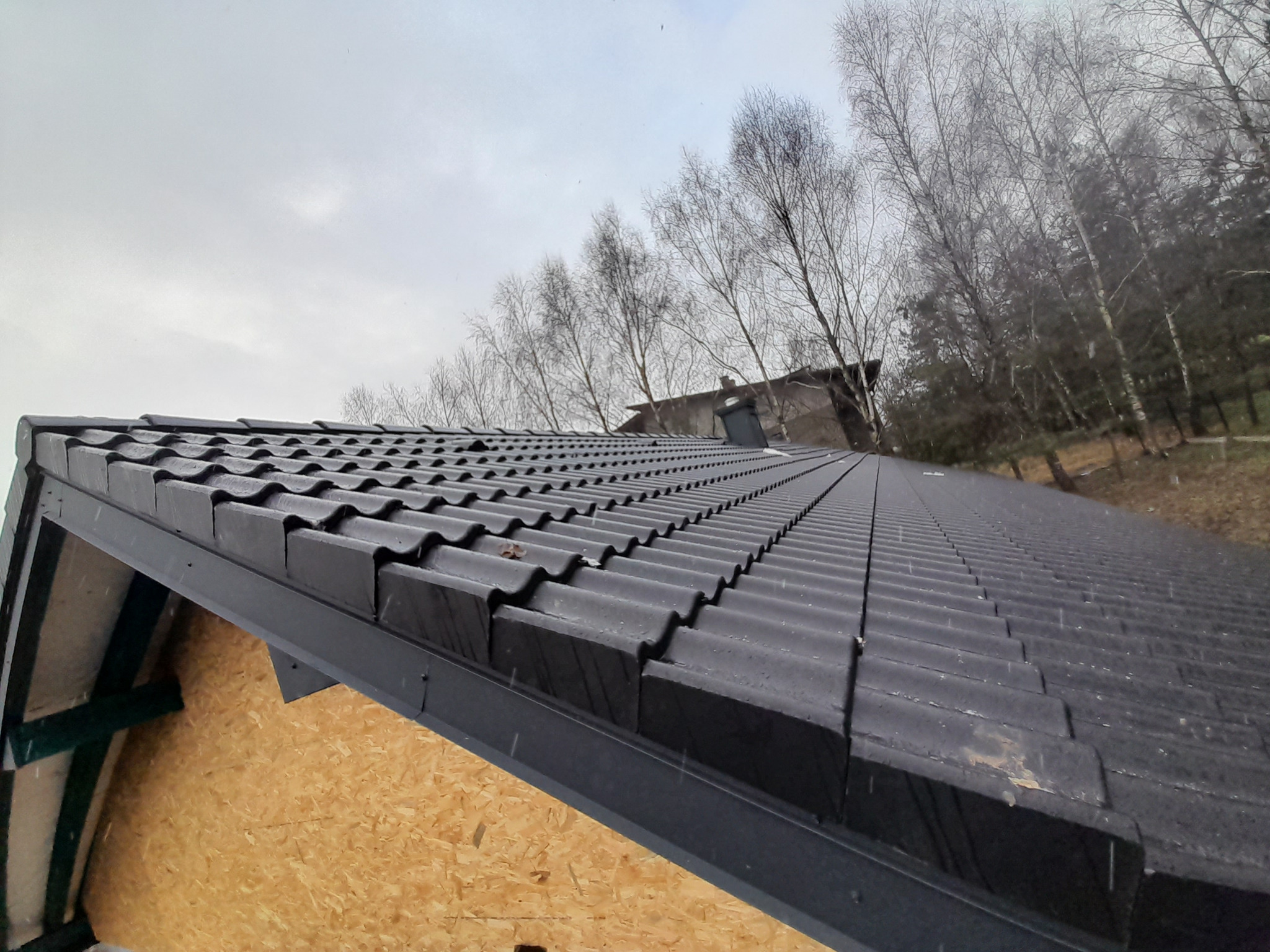 Nowy dach z czarnej dachówki ceramicznej ułożony na drewnianej konstrukcji, widok z dołu na tle pochmurnego nieba i drzew bez liści.