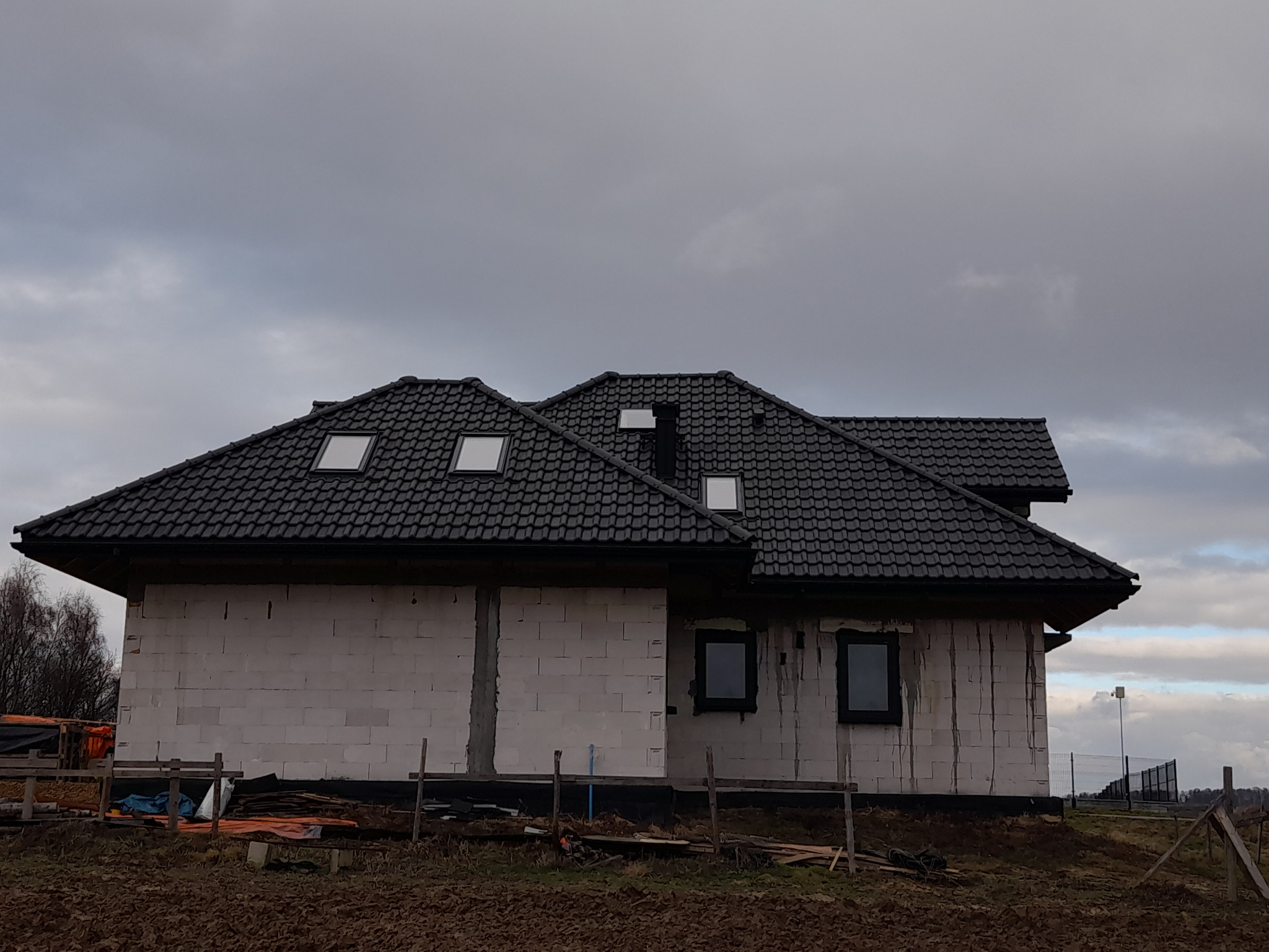 Surowy stan domu jednorodzinnego z ciemną dachówką i oknami dachowymi, ściany z białego bloczka, widoczne ślady po zaprawie, teren wokół domu niezagospodarowany, pochmurne niebo.