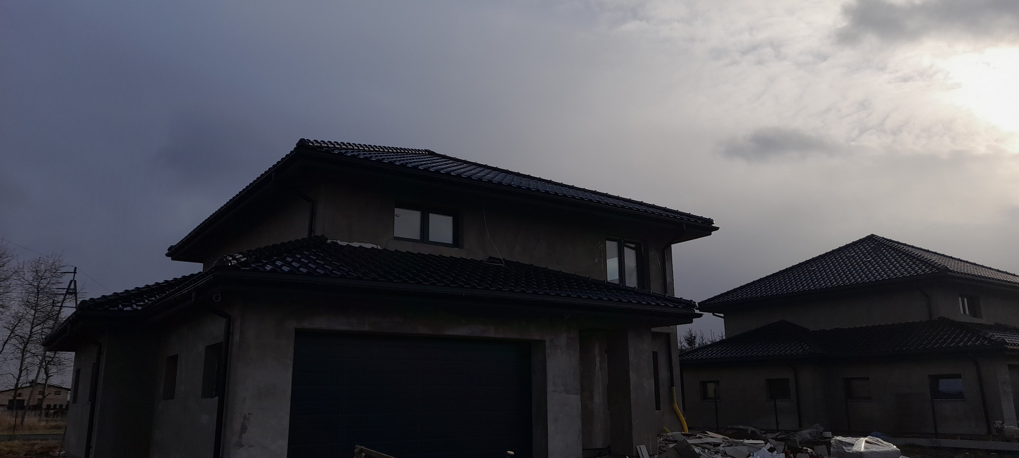 Dwa domy w stanie surowym zamkniętym z czarną dachówką karpiówką, widoczne orynnowanie, elewacja szara, pochmurne niebo.