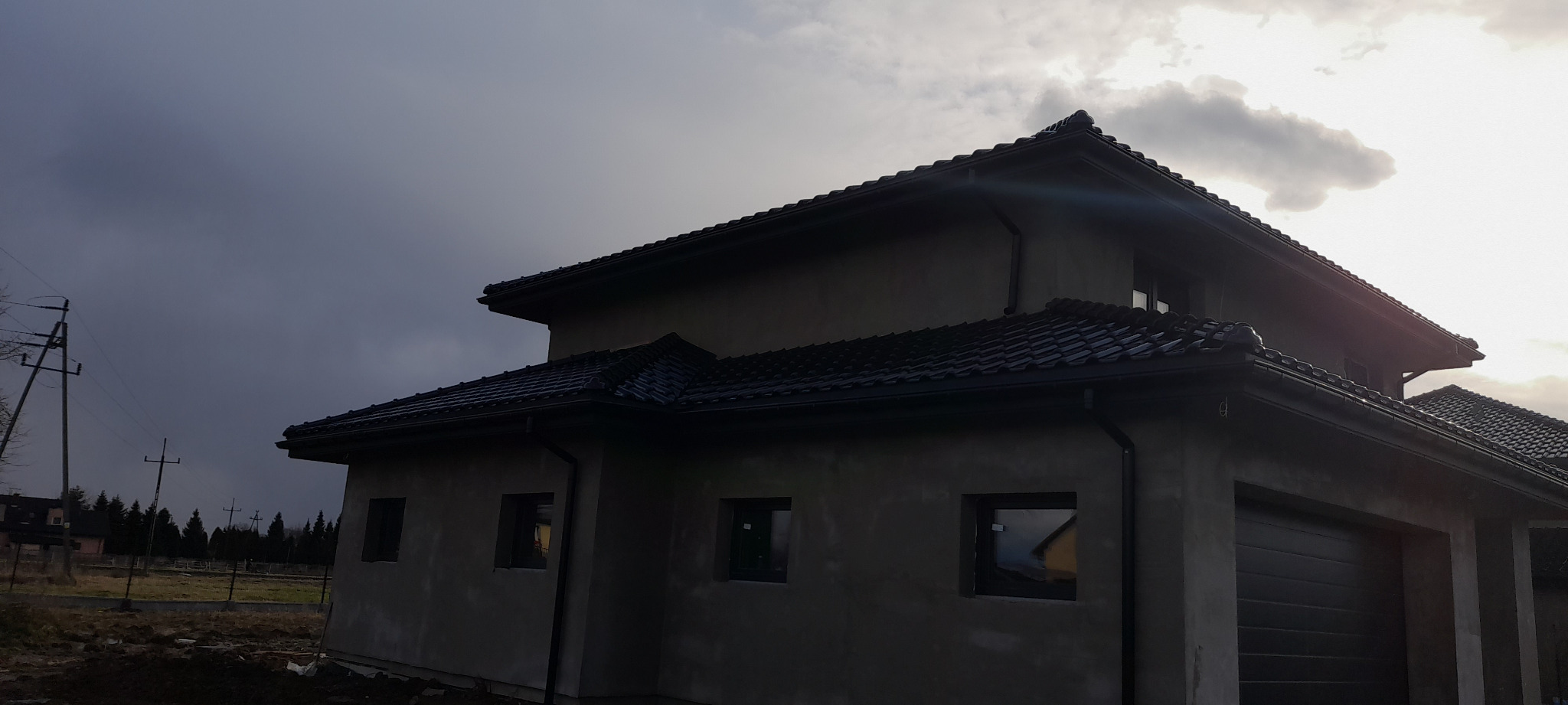 Nowoczesny dom jednorodzinny w stanie surowym zamkniętym z czarną dachówką ceramiczną, widoczne orynnowanie i obróbki blacharskie, na tle pochmurnego nieba.