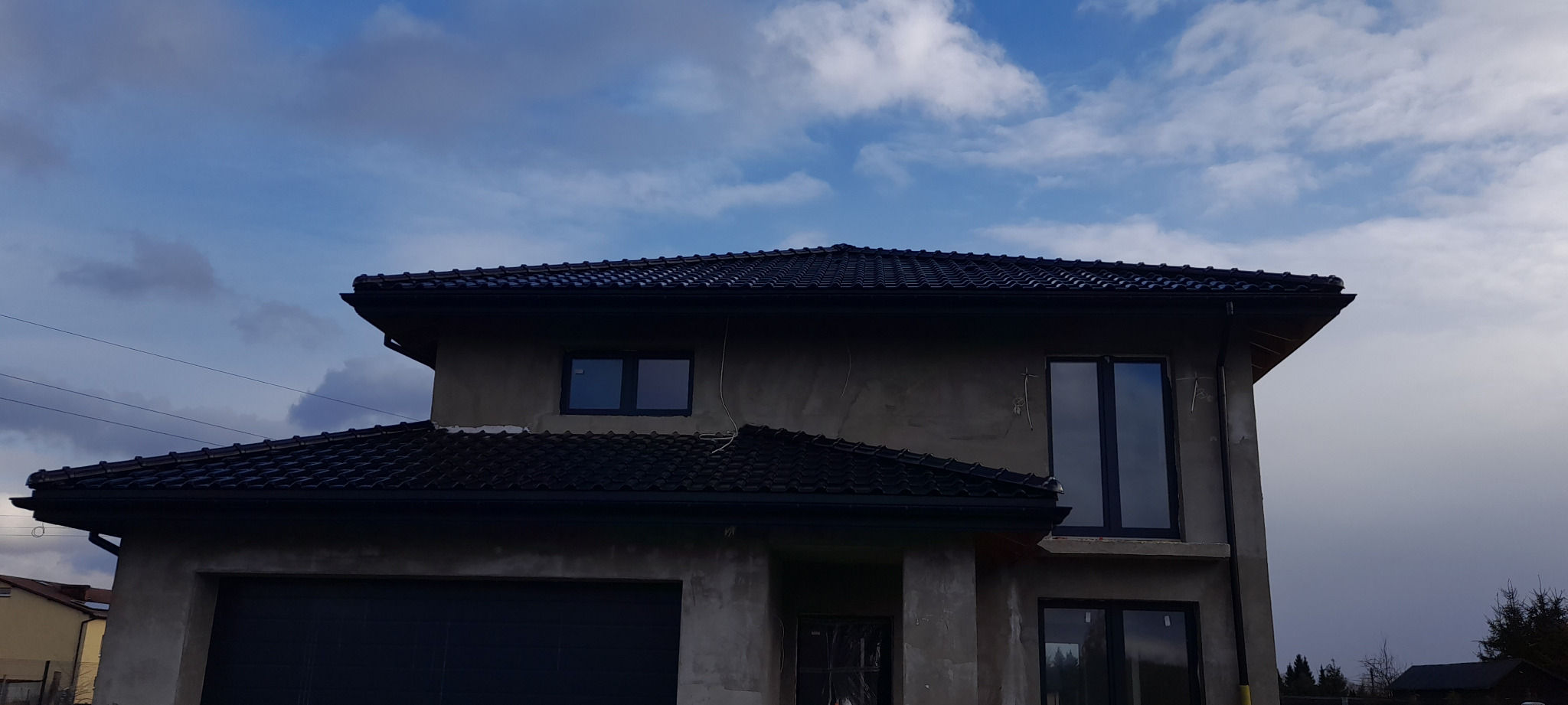 Nowoczesny dom w trakcie budowy z ciemną dachówką i rynnami, widok na fragment elewacji i niebo z chmurami.