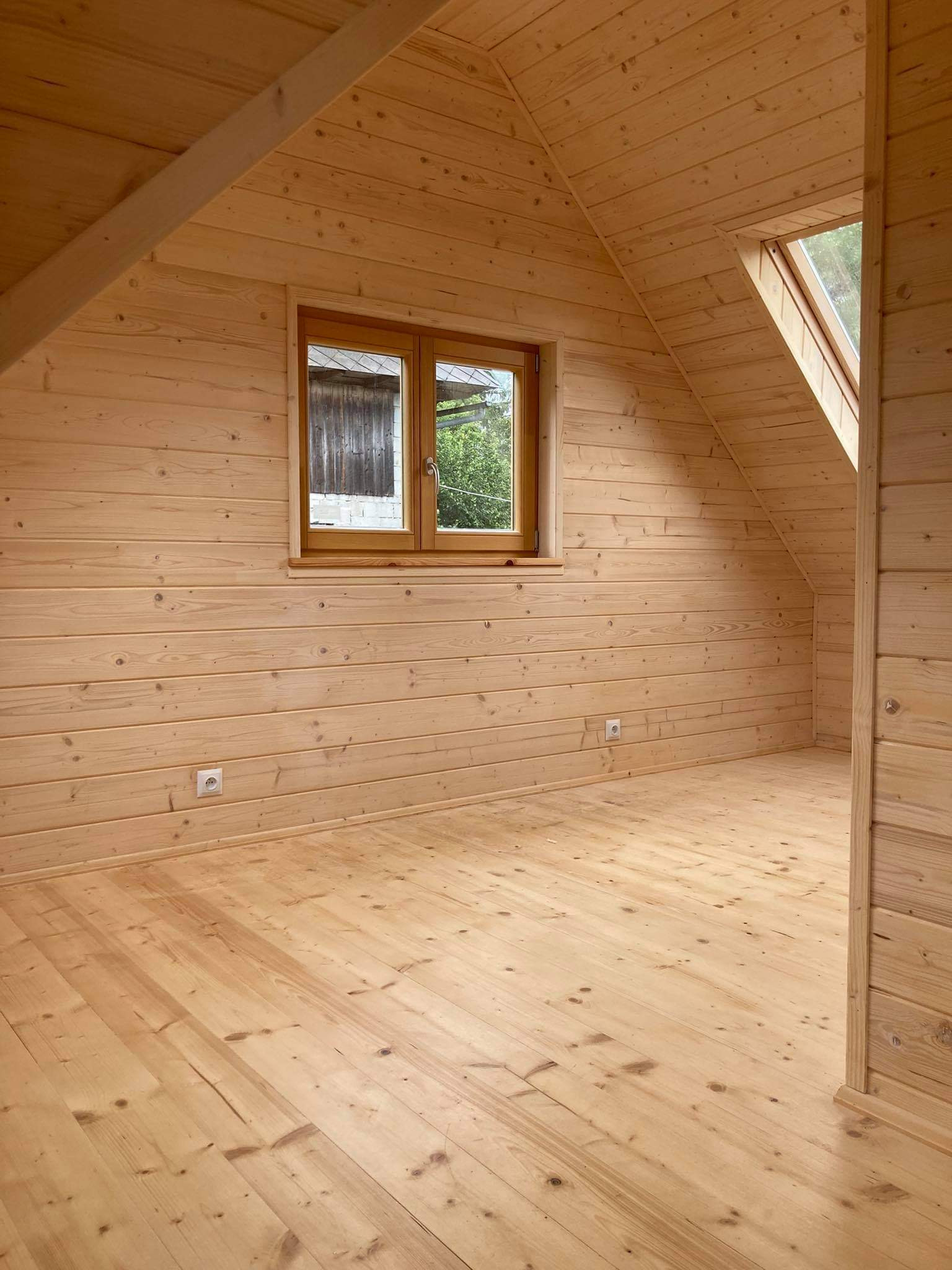 Wnętrze poddasza wykończonego boazerią z jasnego drewna, widoczne okno dachowe i okno szczytowe z widokiem na zielony ogród.