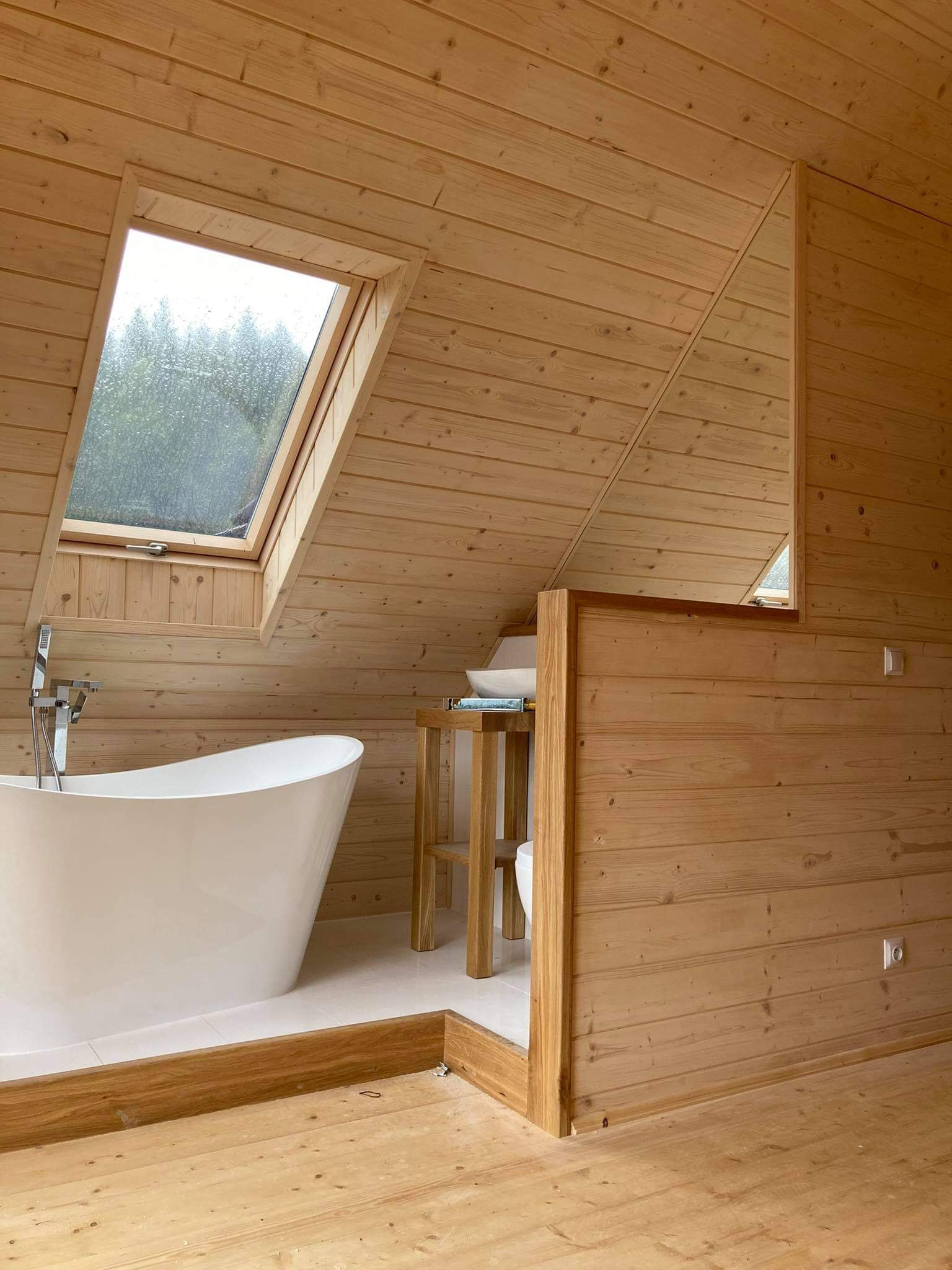 Jasne drewniane wnętrze łazienki na poddaszu z oknem dachowym, białą wanną wolnostojącą i umywalką na drewnianym stoliku.