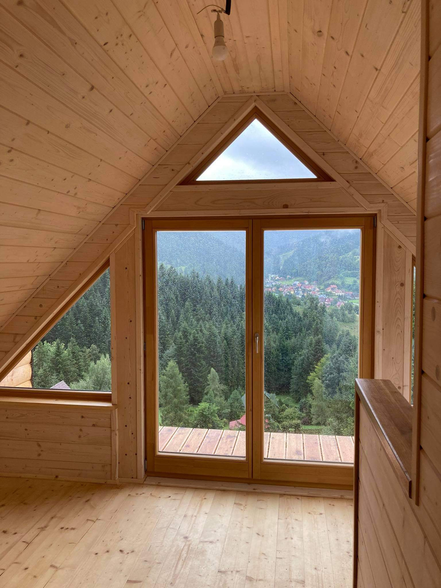 Wnętrze poddasza wykończonego boazerią, z widokiem na las i zabudowę przez duże okno z trójkątnym świetlikiem.
