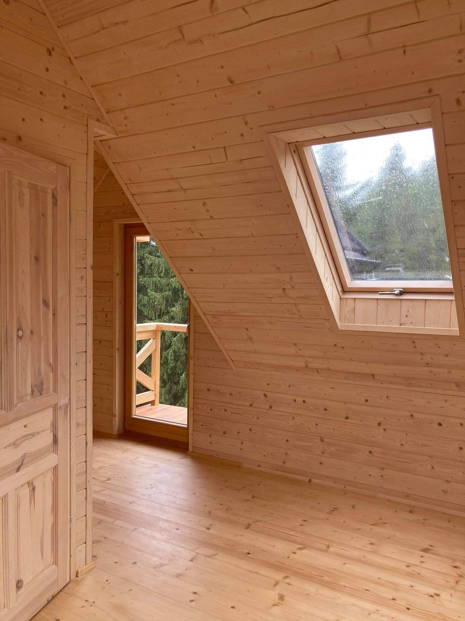 Wnętrze poddasza wykończonego boazerią z oknem dachowym i wyjściem na balkon, widok na zewnątrz z dominującą zielenią drzew.