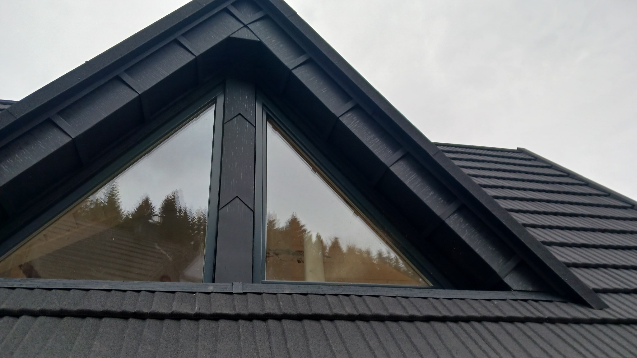 Szczegółowe ujęcie dachu z czarnej dachówki ceramicznej i trójkątnego okna, w którym odbijają się drzewa