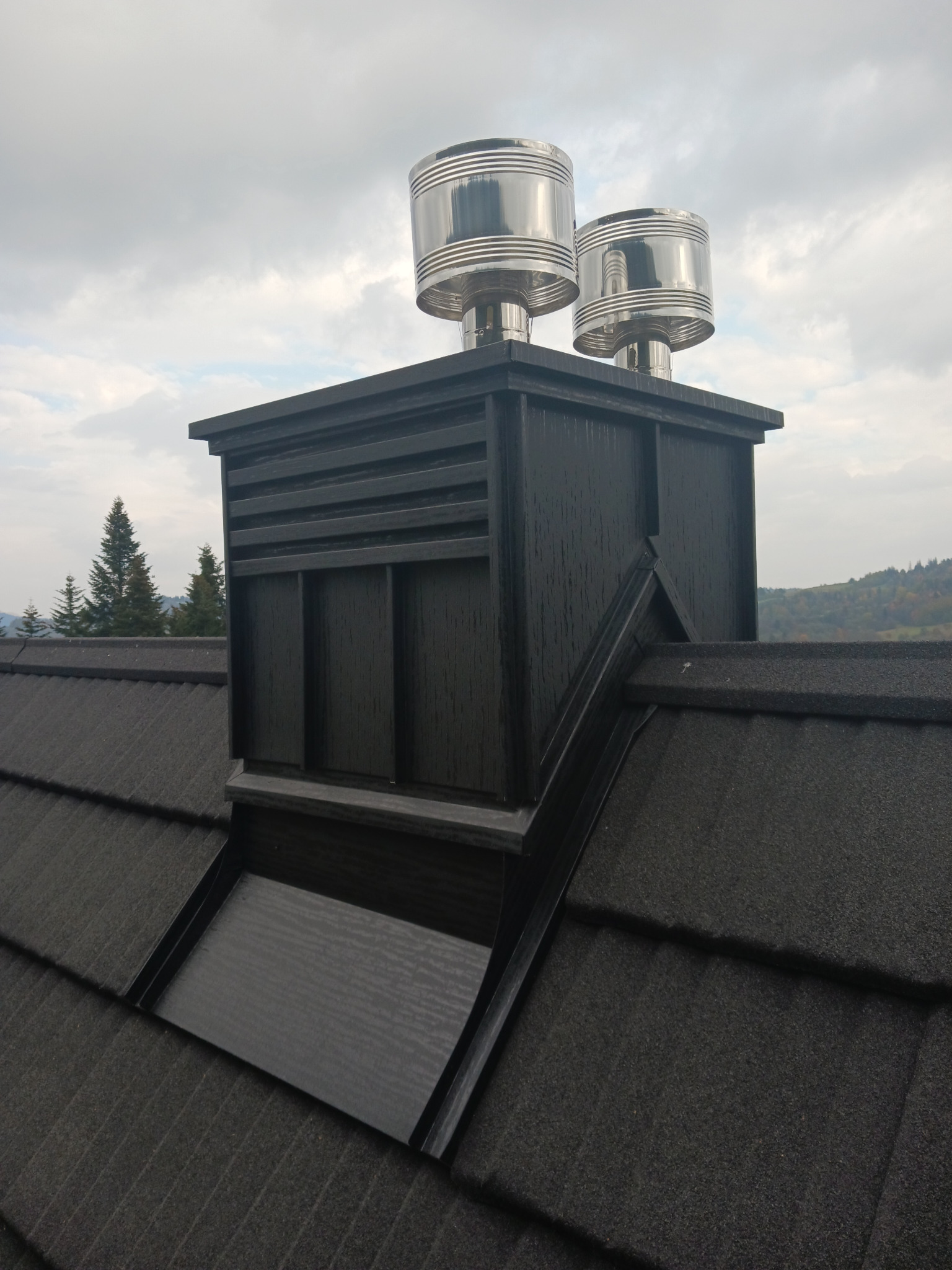 Czarny komin z dwoma metalowymi nasadami wentylacyjnymi na dachu z bitumicznej dachówki, widok na zalesione wzgórza w tle.
