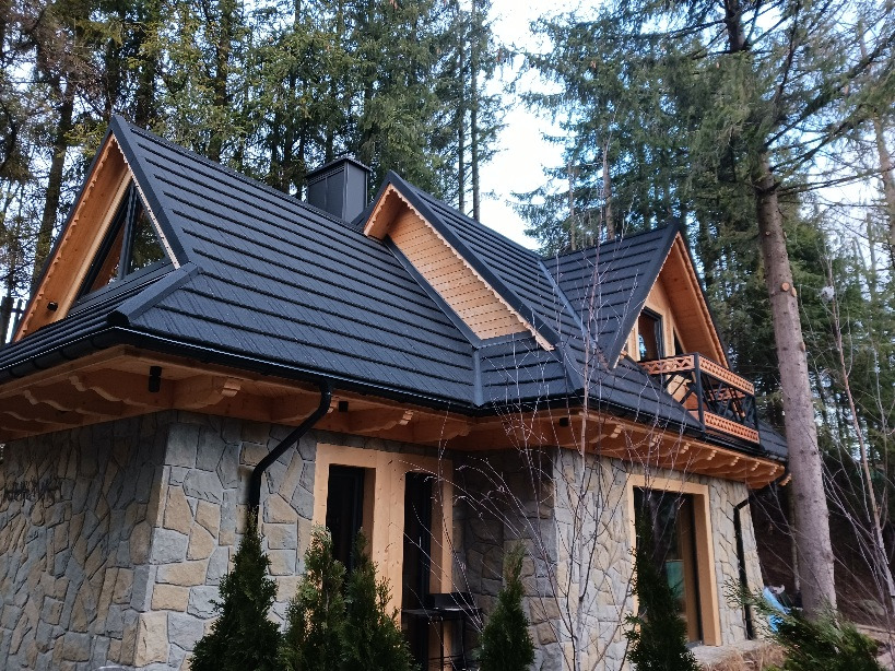 Dom w stylu góralskim z czarnym dachem z blachodachówki, kamienną elewacją i drewnianą więźbą dachową, otoczony drzewami.