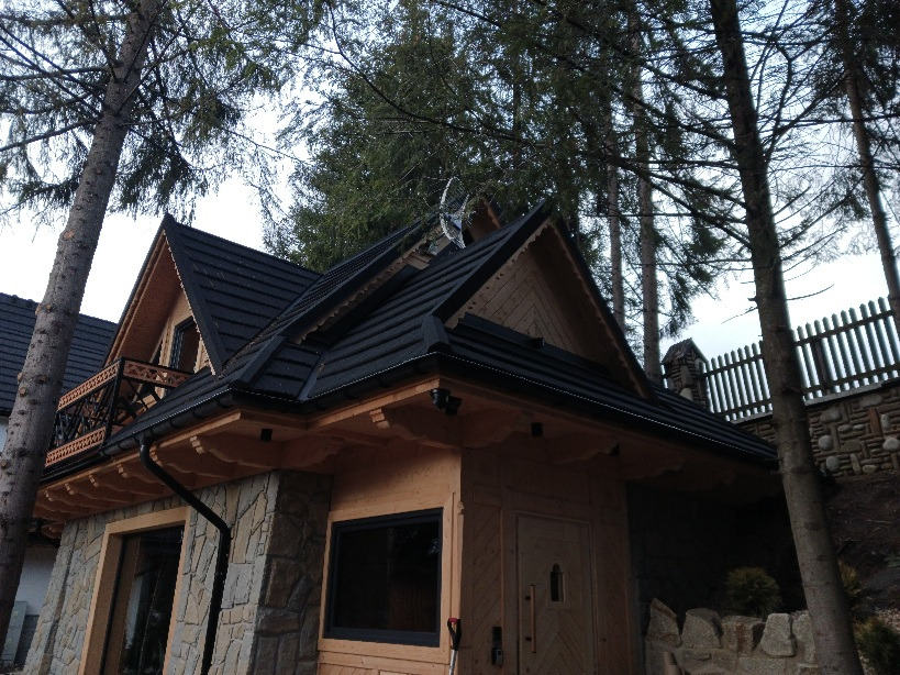 Dom w stylu góralskim z czarnym dachem z blachy na rąbek stojący, drewnianymi elementami wykończeniowymi i małym balkonem z ozdobną balustradą, widoczny z dołu na tle drzew.