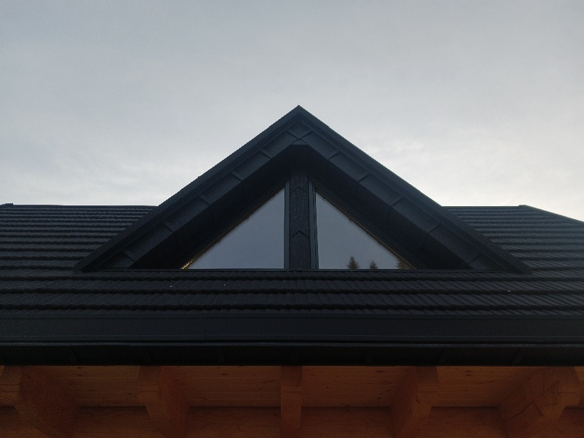 Ciemny, skośny dach z oknem trójkątnym na poddaszu, widoczne drewniane elementy konstrukcyjne budynku.