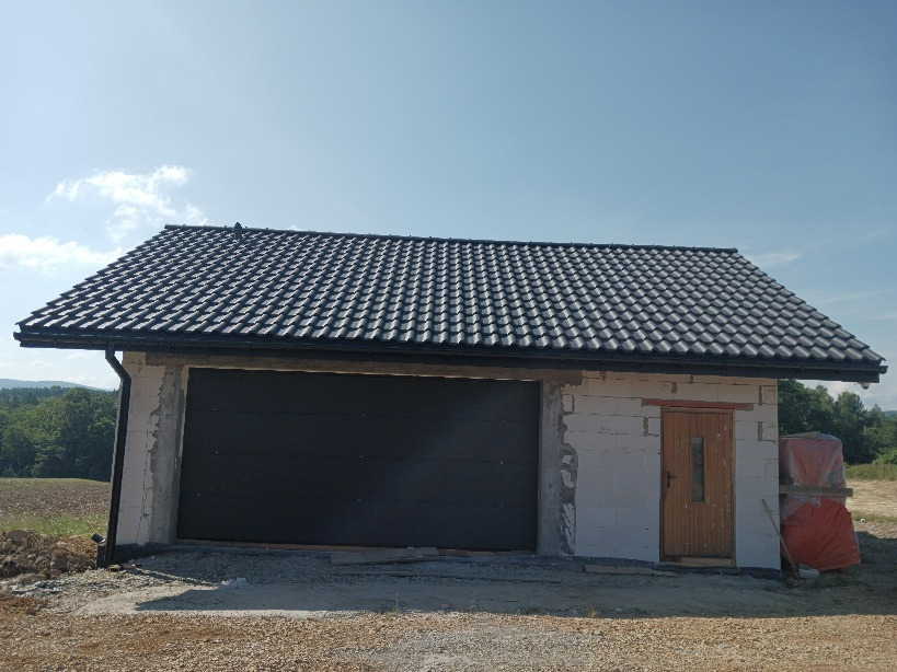 Nowy, ciemny dach z dachówki ceramicznej na garażu z bramą segmentową, widoczne rynny, budynek w stanie surowym otwartym.