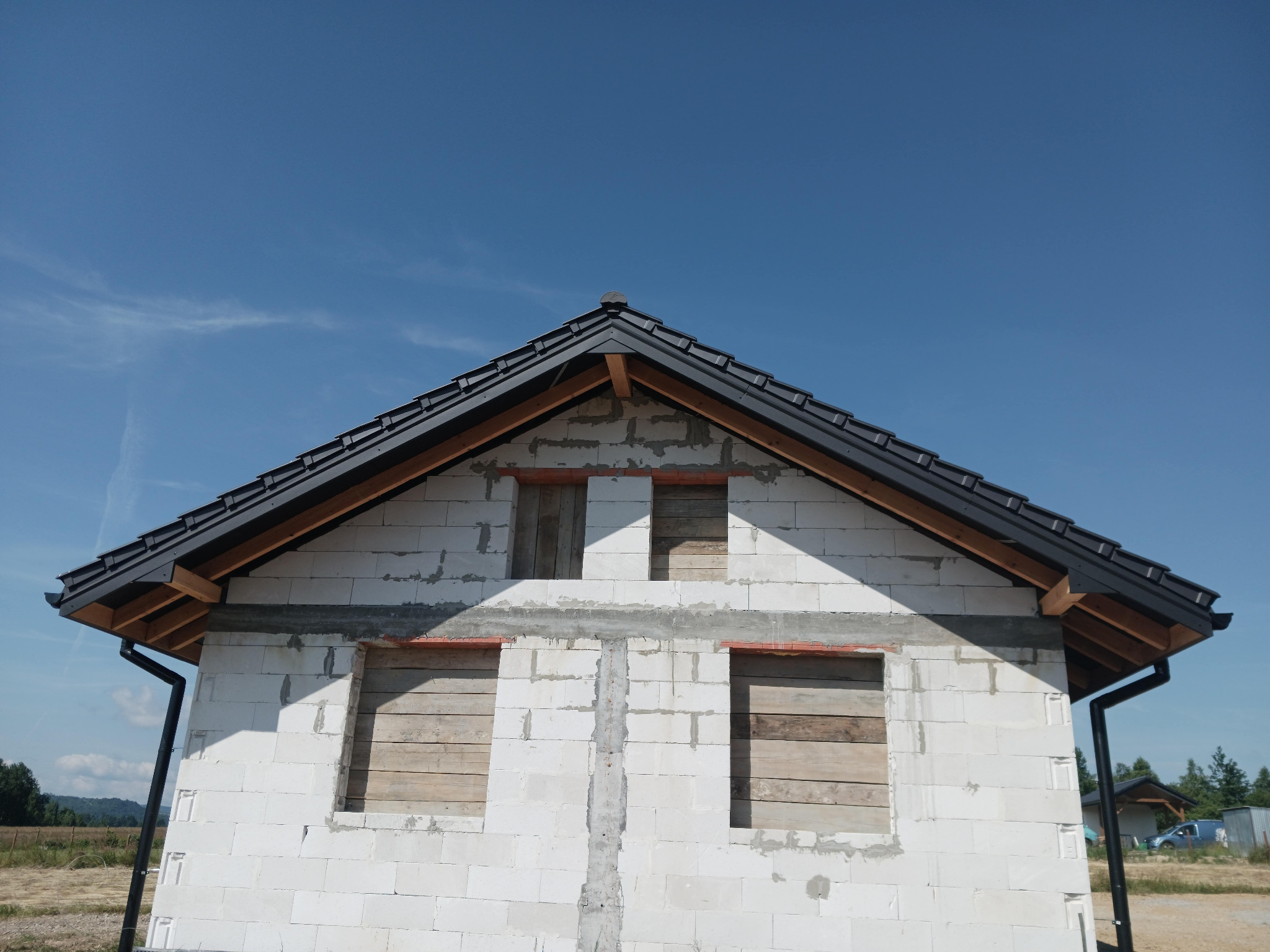 Surowy stan budynku z białych bloczków z zamontowaną czarną blachodachówką i rynnami, okna zabite deskami, widok na błękitne niebo.