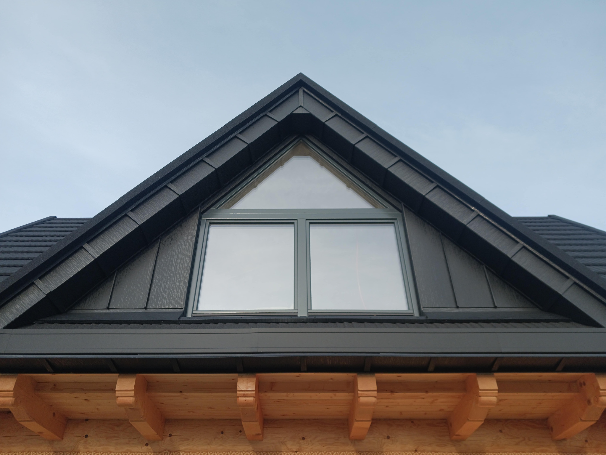 Nowoczesny dach z czarnej blachy trapezowej z trójkątnym oknem na szczycie, drewniane elementy konstrukcyjne widoczne poniżej.
