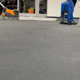 Dwie osoby czyszczą dywan w biurze za pomocą profesjonalnych maszyn: jedna szoruje, a druga odsysa wodę. Widoczny efekt czyszczenia na fragmencie dywanu.