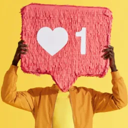 Osoba trzyma przed twarzą różową piniatę w kształcie dymka z ikoną serca i cyfrą 1, symbolizującą polubienie w mediach społecznościowych na jaskrawożółtym tle.