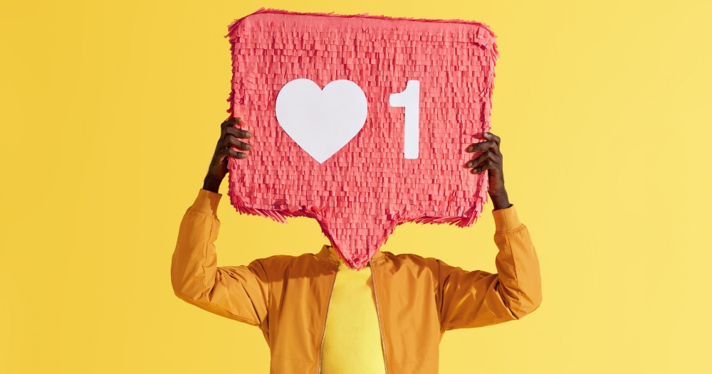 Osoba trzyma przed twarzą różową piniatę w kształcie dymka z ikoną serca i cyfrą 1, symbolizującą polubienie w mediach społecznościowych na jaskrawożółtym tle.