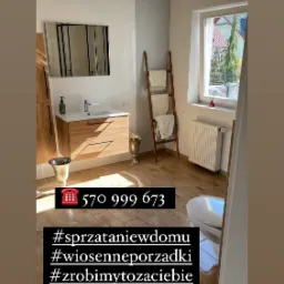 Wnętrze łazienki po sprzątaniu: drewnopodobne płytki, umywalka z szafką, lustro, drabinka na ręczniki, grzejnik pod oknem. Widoczne detale jak złote pojemniki na akcesoria.