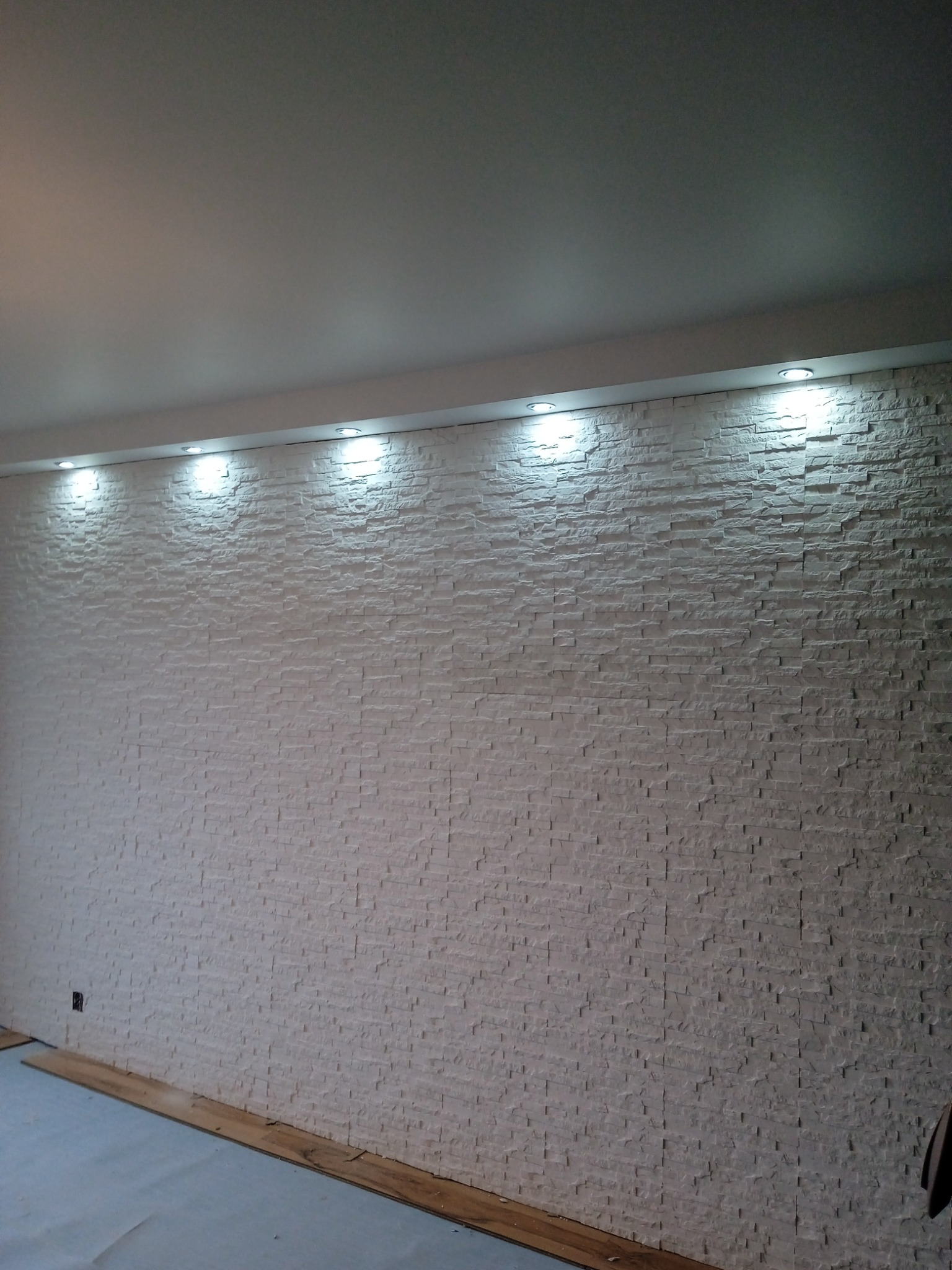 Ściana wewnętrzna wykończona białym kamieniem dekoracyjnym, oświetlona punktowo od góry, widoczne fragmenty podłogi w trakcie układania paneli.