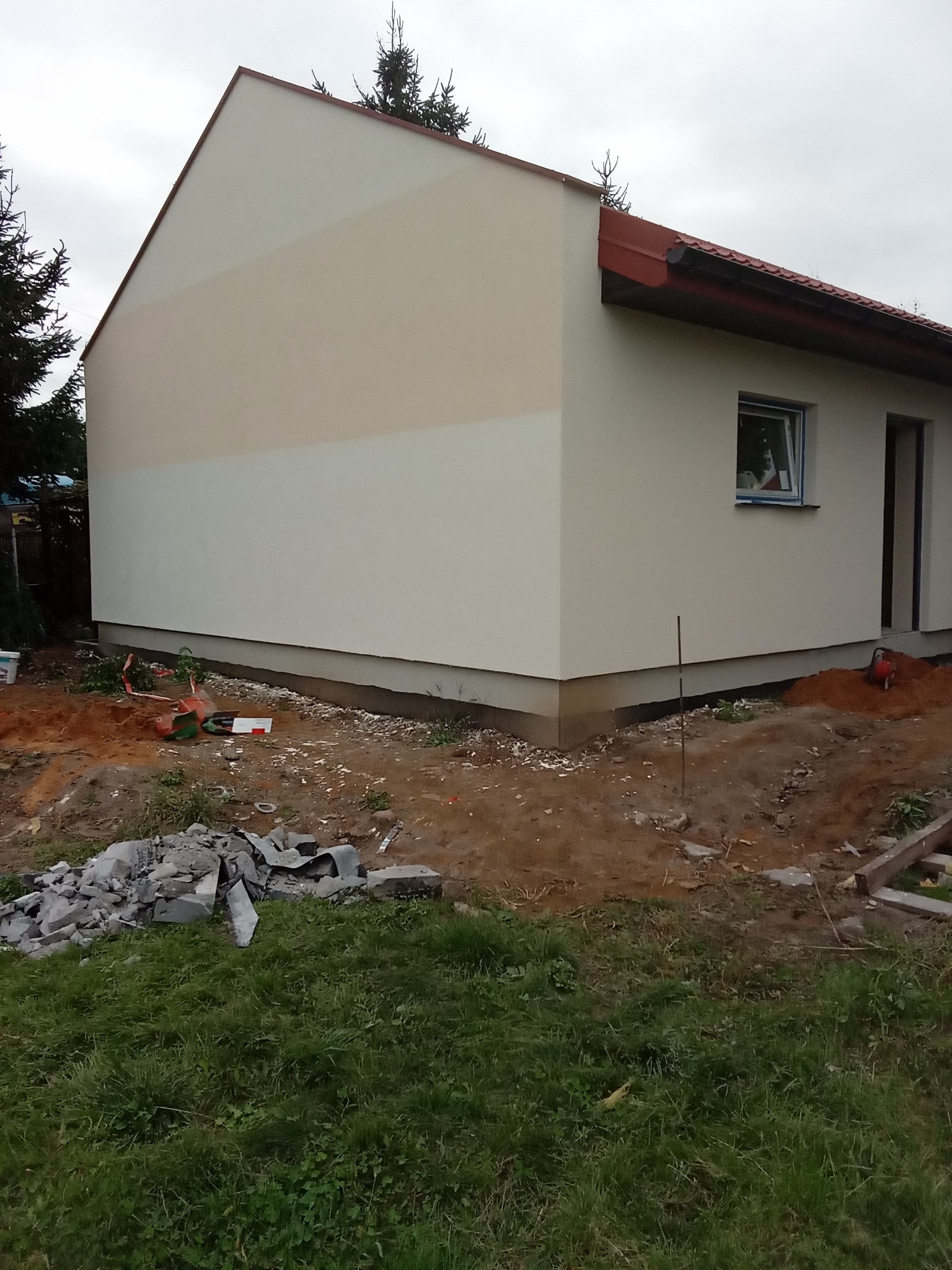 Fragment ocieplonej elewacji domu jednorodzinnego z widocznymi różnicami w odcieniach farby, gruzem i narzędziami na trawniku.