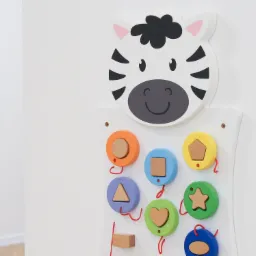 Drewniana zabawka edukacyjna w kształcie zebry z kolorowymi kółkami i drewnianymi figurami geometrycznymi przymocowanymi sznurkami, obok widoczna zabawka z przesuwankami, na białej ścianie.