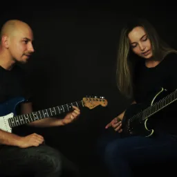 Mężczyzna z niebieską gitarą Fender uczy kobietę grać na czarnej gitarze elektrycznej na ciemnym tle.