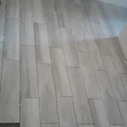 Układanie podłogi z płytek imitujących drewno w odcieniach szarości, widoczne krzyżyki dystansowe i kątownik monterski.