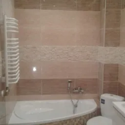 Jasna łazienka z narożną wanną obudowaną mozaiką, beżowe płytki na ścianach z poziomym pasem dekoracyjnym, biała drabinka na ręczniki i podwieszany sufit z oświetleniem punktowym.