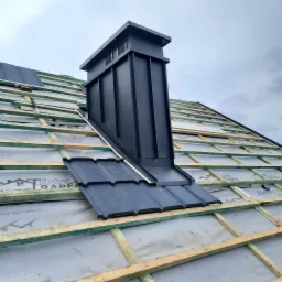 Montaż czarnego komina wentylacyjnego na dachu z blachy trapezowej, widoczne drewniane łaty i membrana dachowa.